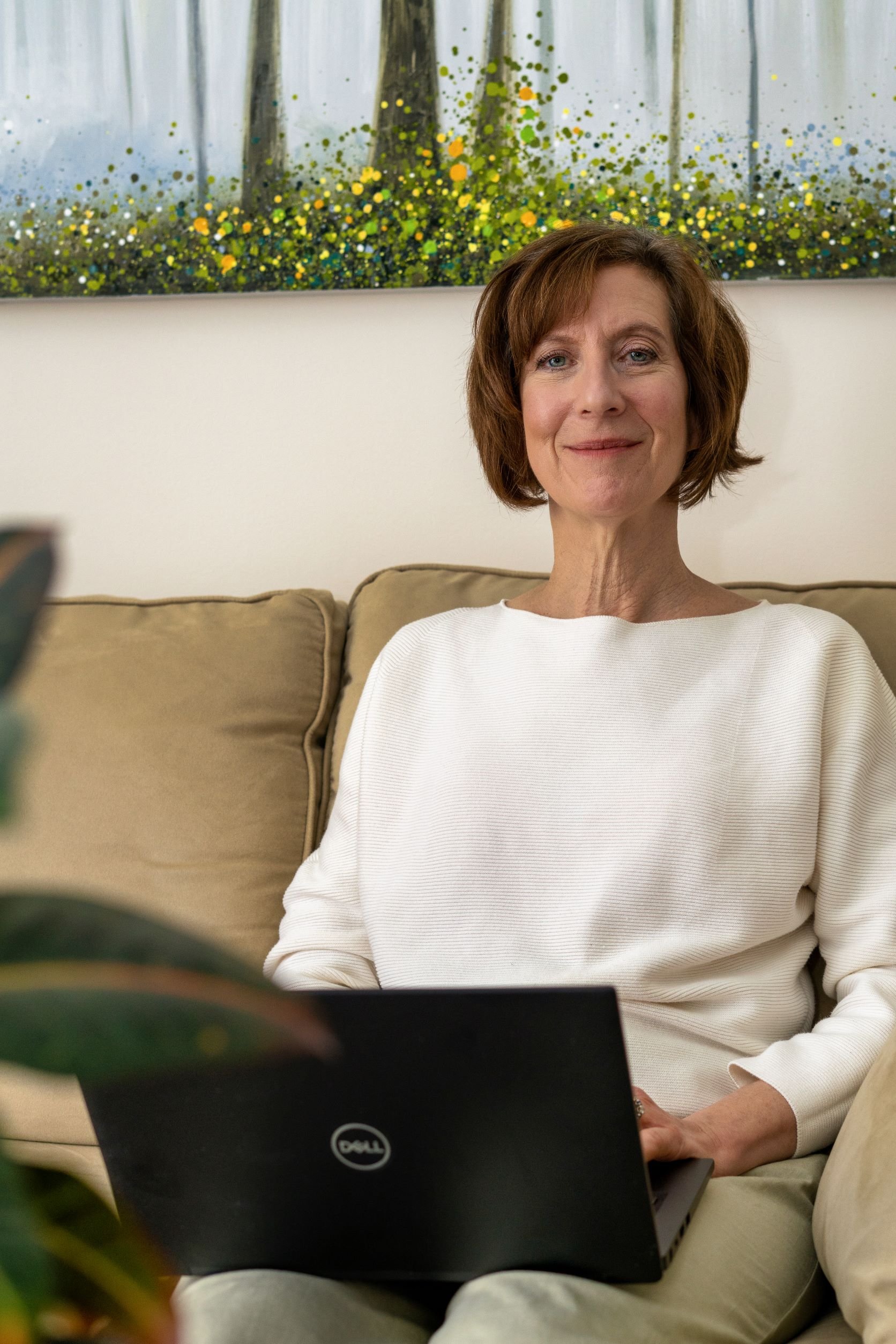 A woman with short brown hair and a white top sitting on a beige couch with a Dell laptop on her lap, in front of a colorful abstract painting with green, yellow, and black splashes.