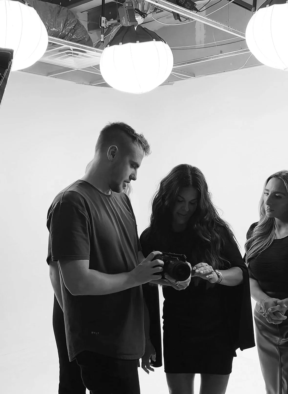Three people in a photo studio look at a camera held by a young man, with studio lighting and equipment overhead.