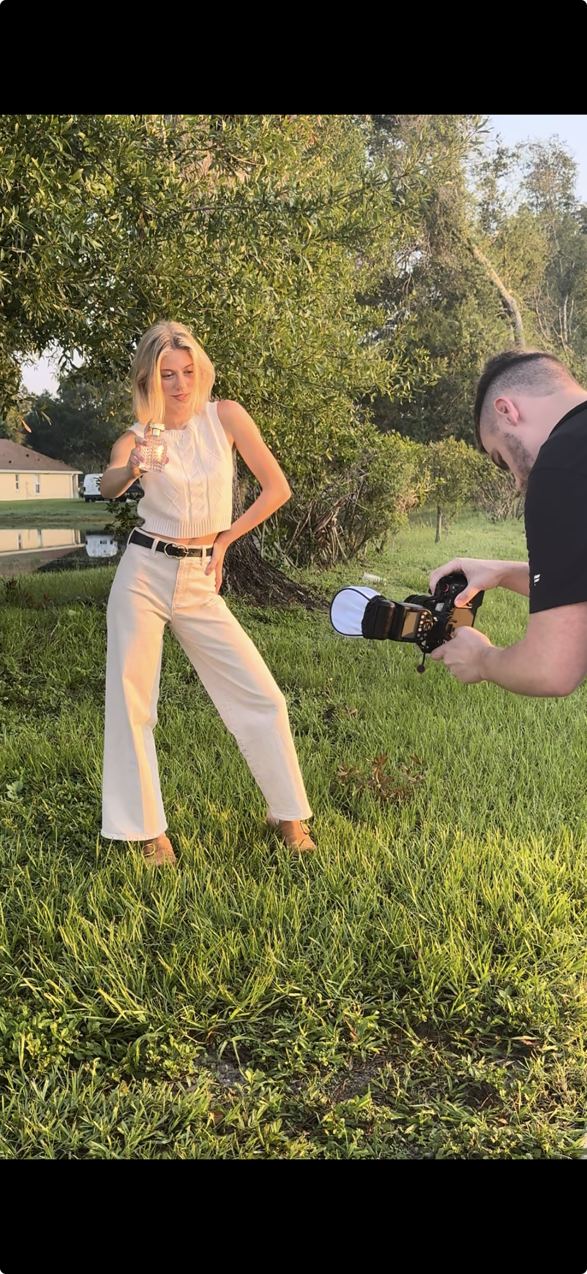 A woman standing on green grass, holding a glass in her right hand, while a man takes her photo with a camera outdoors during sunset, with trees and a house in the background.