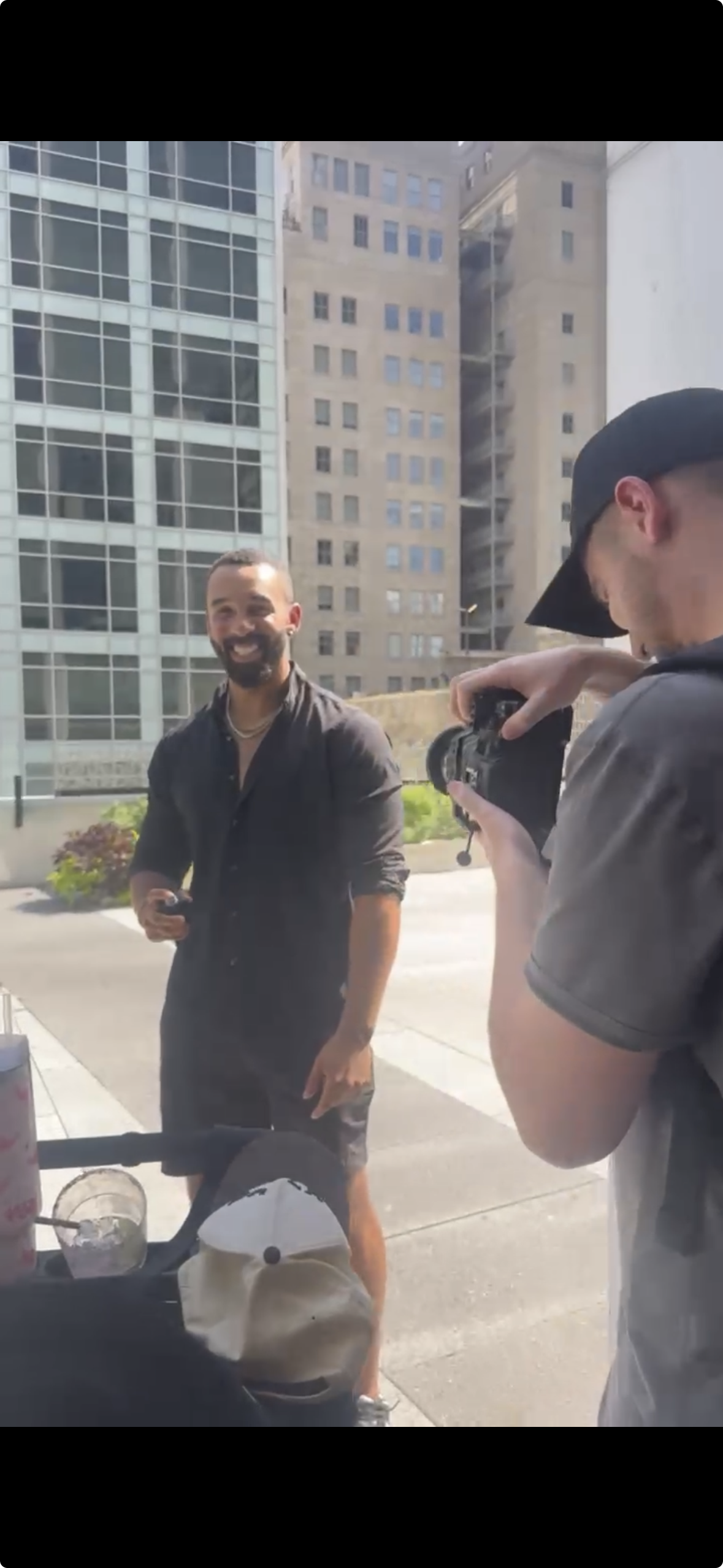 A man with a beard smiling and holding a phone stands outside in an urban setting, while a person with a camera photographs him. Tall buildings are visible in the background.