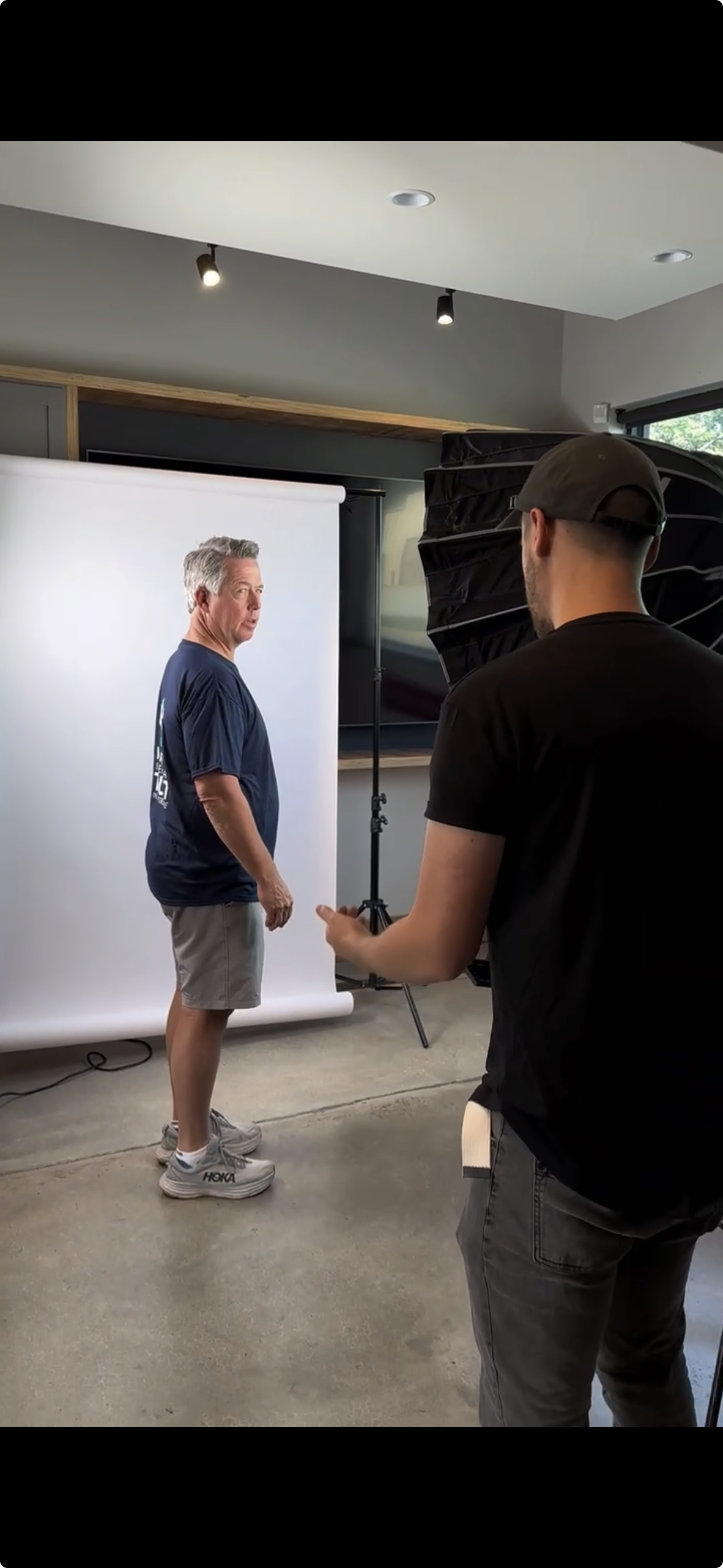 A man in a blue shirt and gray shorts is standing in front of a white backdrop in a photography studio. He is looking over his shoulder. Another person, wearing a black shirt and gray pants, is operating a piece of photography equipment. The studio h
