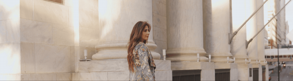 A woman with long brown hair in a floral jacket standing near large marble pillars on a city street.