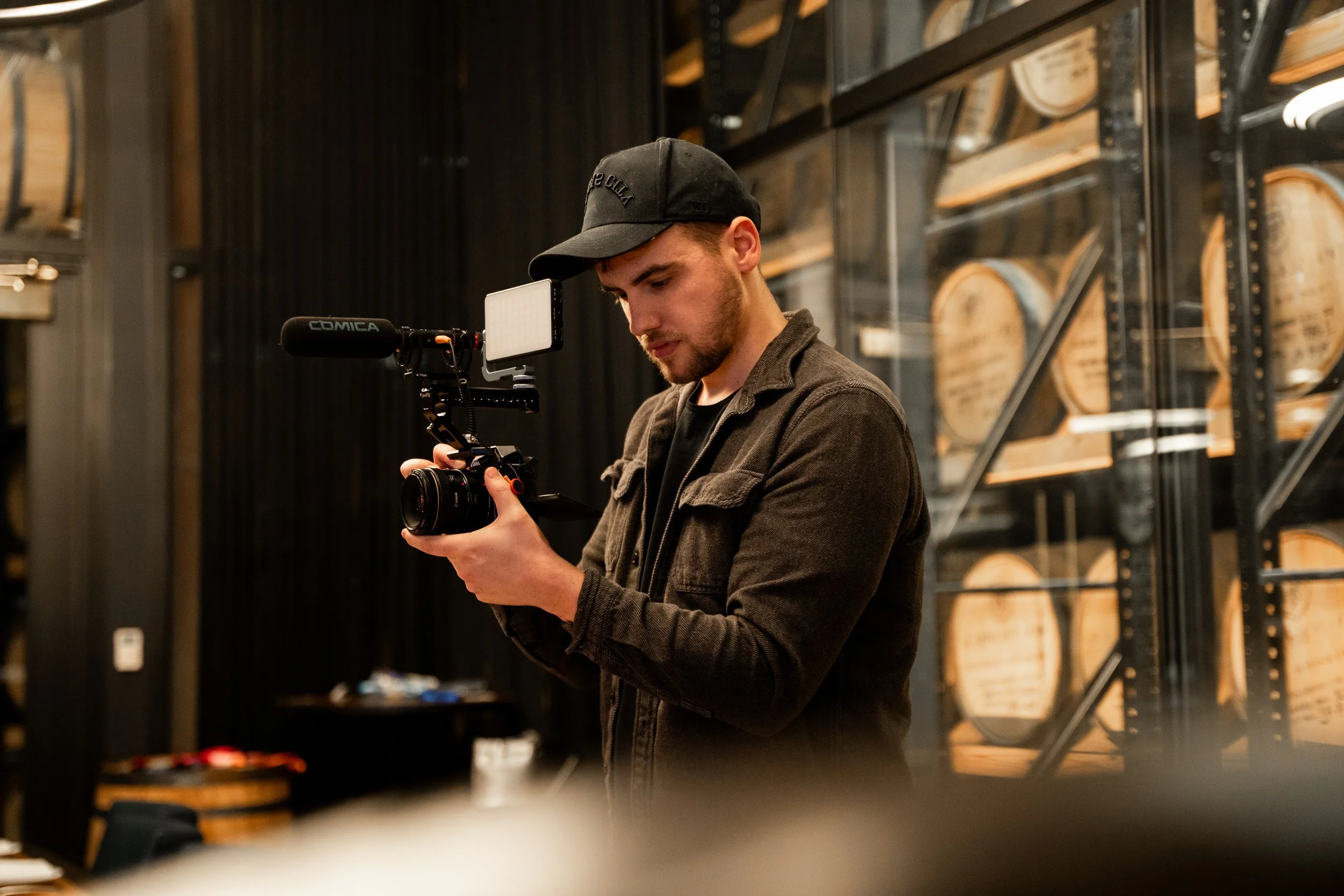 A man filming with a camera rig in a room with shelves of wooden barrels.