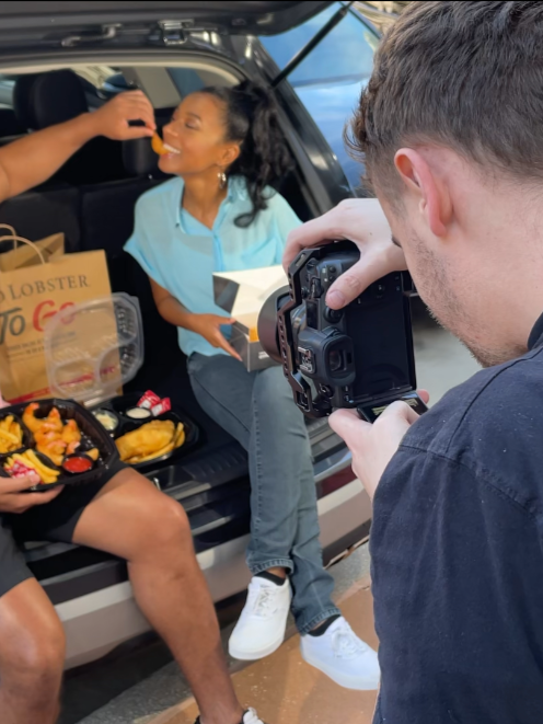A woman sitting on the open trunk of a car, enjoying food from a takeout box and a funnel cake, while a person beside her is feeding her with fries. A photographer in the foreground is taking a picture of the woman.