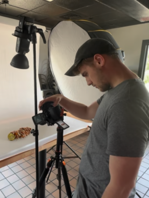 A man adjusting a professional camera on a tripod in a photography studio with a large white reflector and an overhead light.