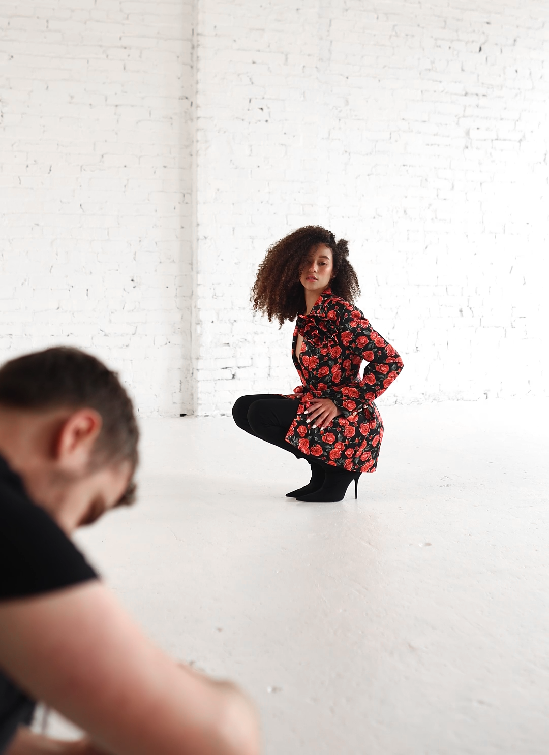 A woman with curly hair squats in front of a white brick wall, wearing a floral coat with red roses and black high-heeled boots. A man is in the foreground, blurred and looking downward.