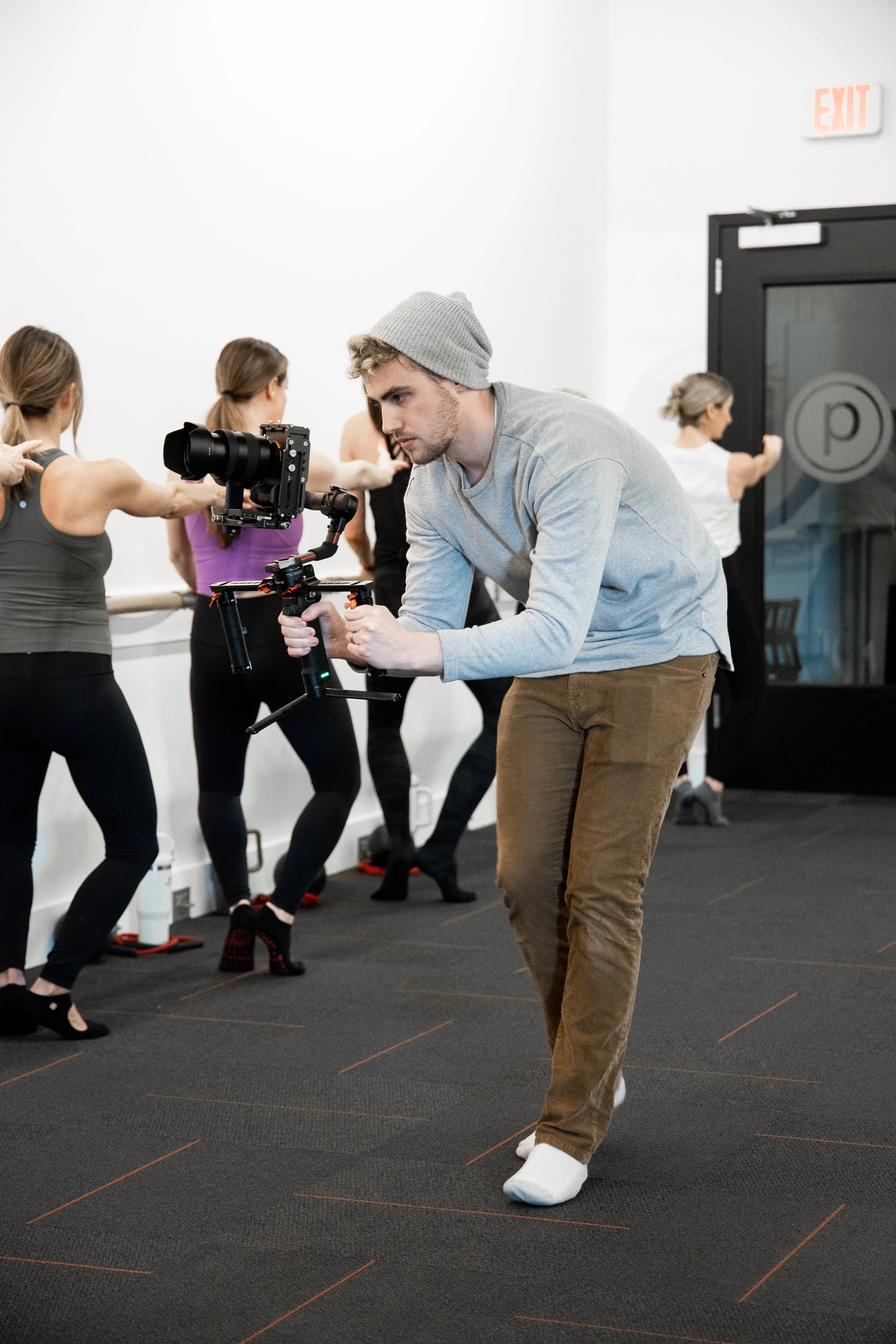 A man in a gray beanie and gray long-sleeve shirt operating a professional camera rig in a dance studio with women stretching and exercising in the background.
