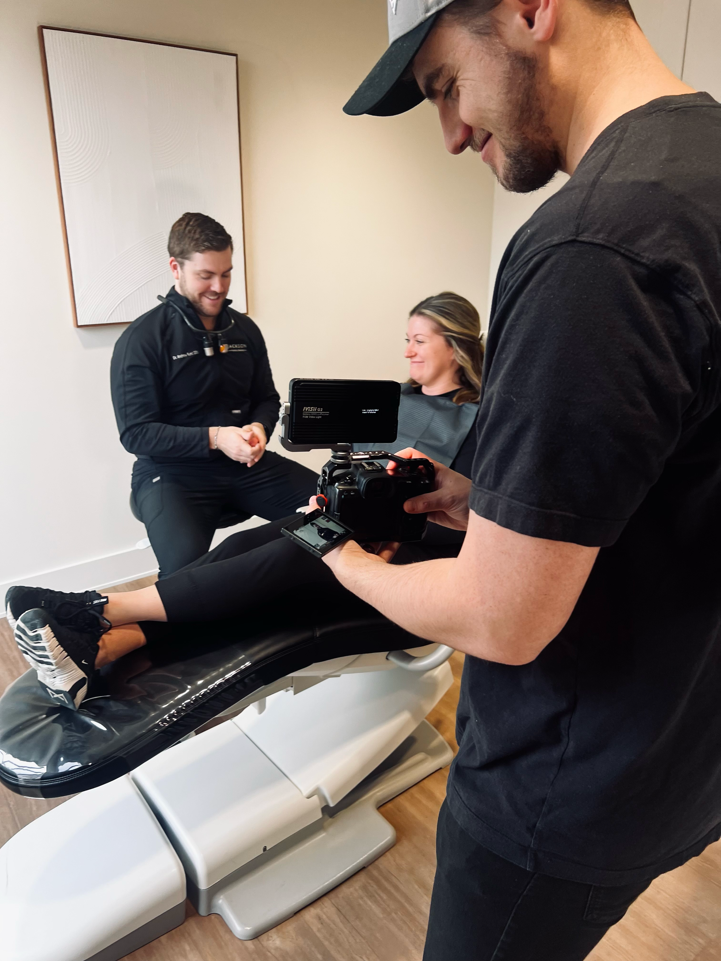 A woman lying on a medical examination table with medical professionals and a photographer. The woman is smiling, the professionals are talking, and the photographer is taking pictures.