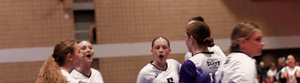 A volleyball team of five young women in white and purple uniforms are huddled together, speaking or shouting during a game or practice in a gymnasium.