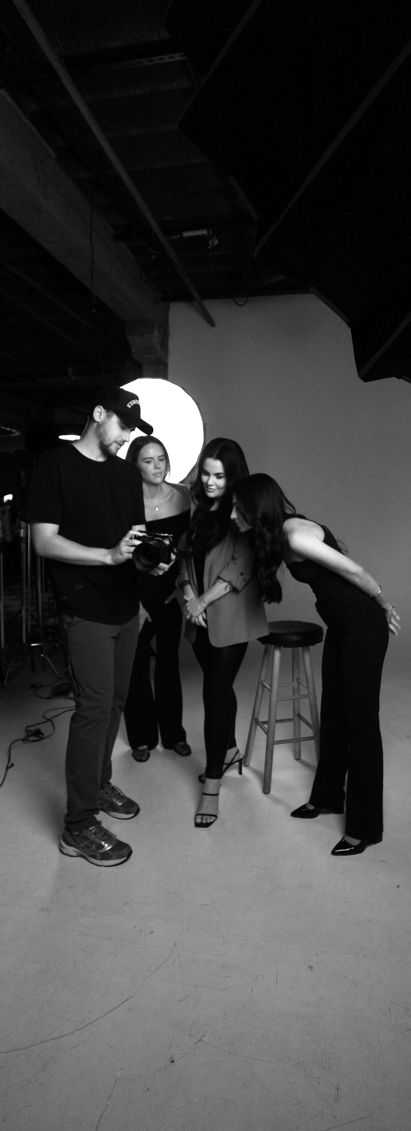 Black and white photo of four people in a photography studio, with three women and one man, two women are sitting and two are standing, while the man shows something on his camera.