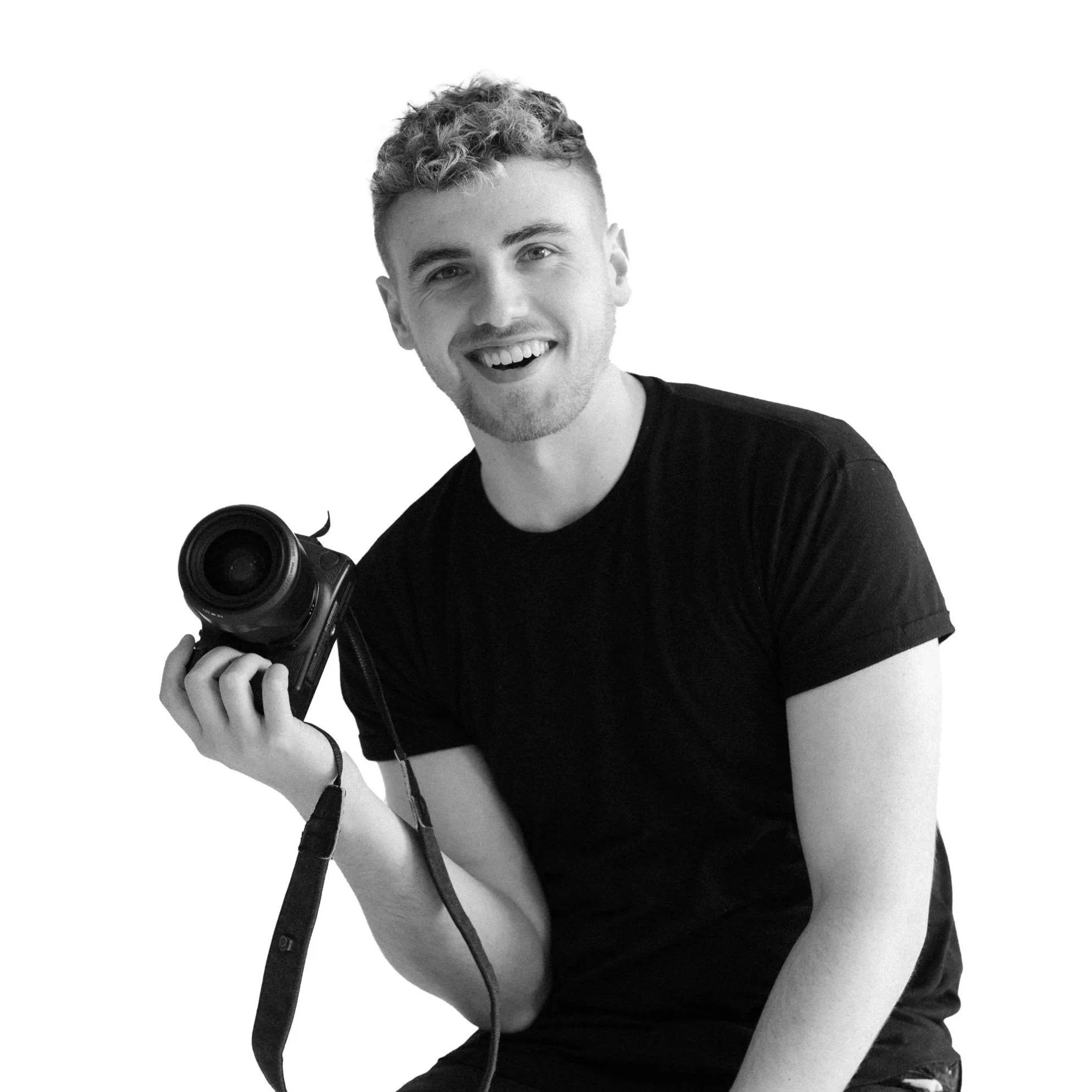 A young man with short, curly hair smiling and holding a camera, wearing a black t-shirt, against a plain white background.