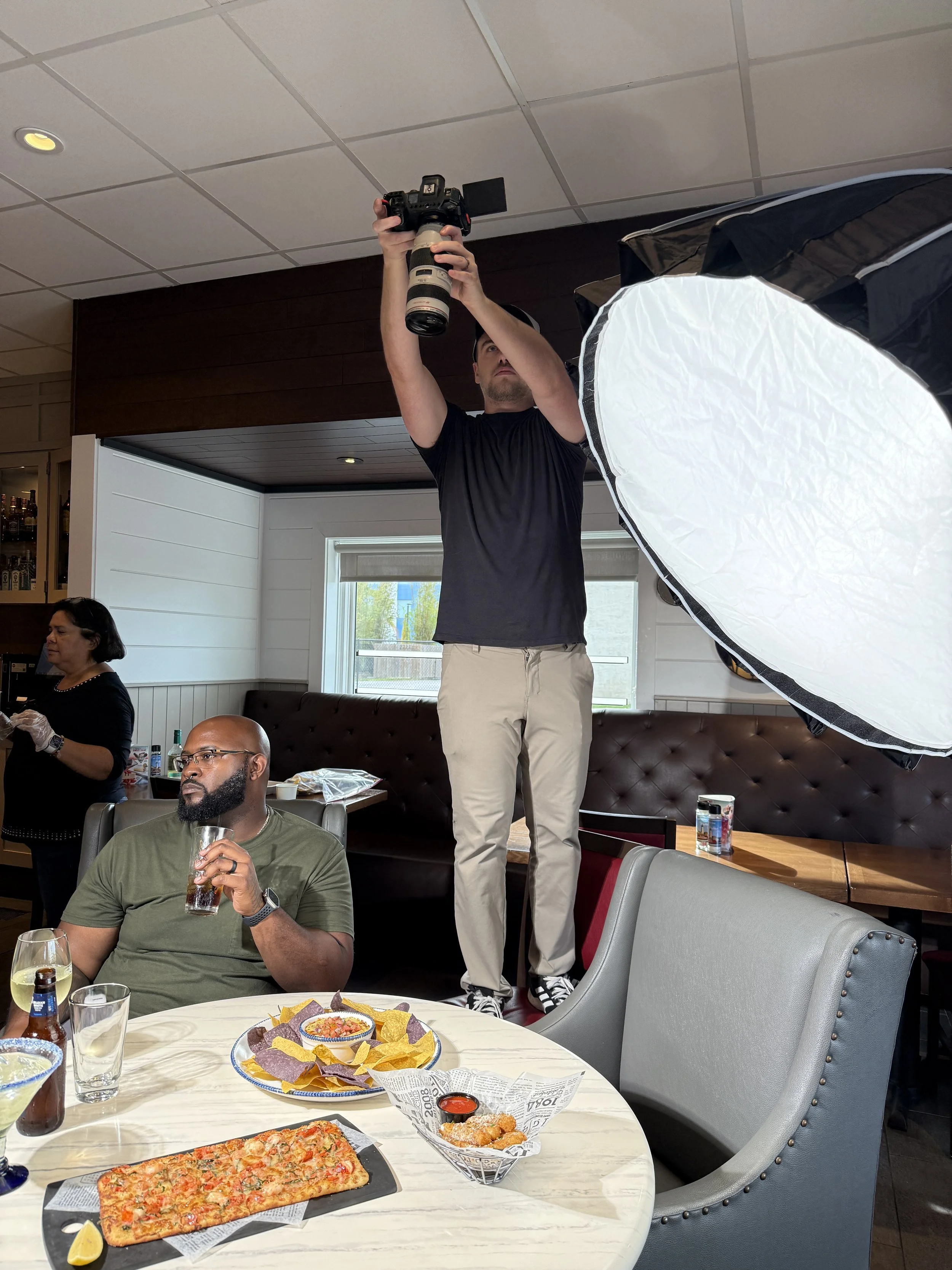 Photographer standing on a chair taking a picture inside a restaurant with people dining at tables.