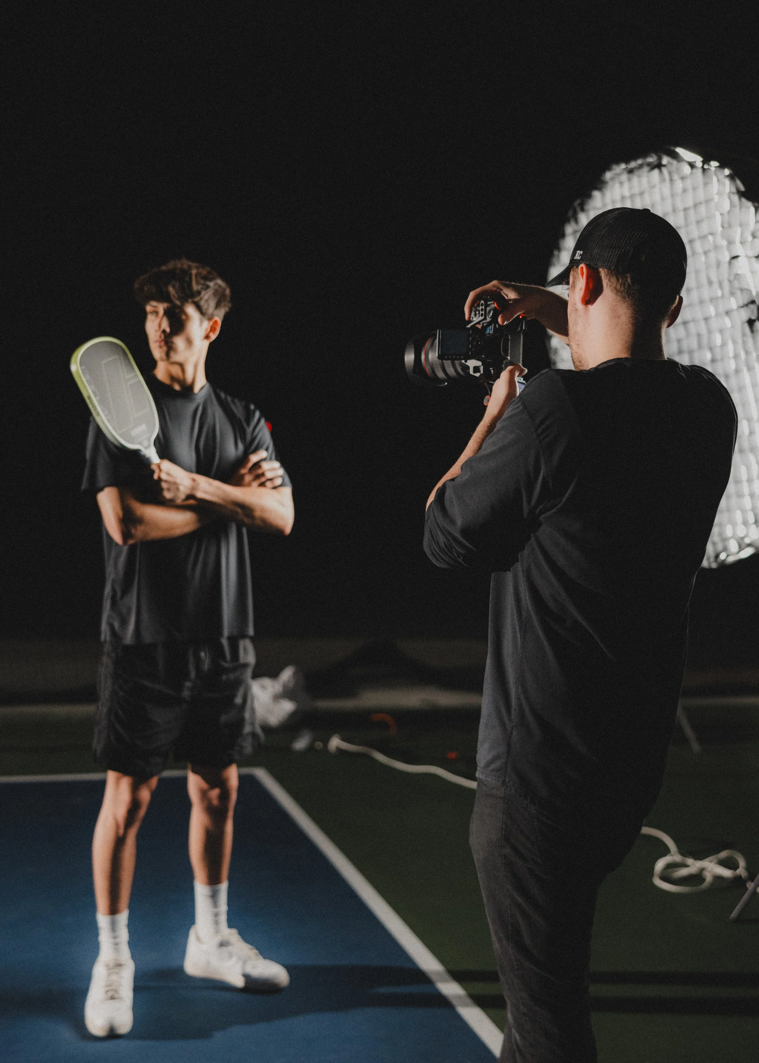 Photographer filming a young male tennis player holding a tennis racket and standing on a court at night, illuminated by a large studio light.