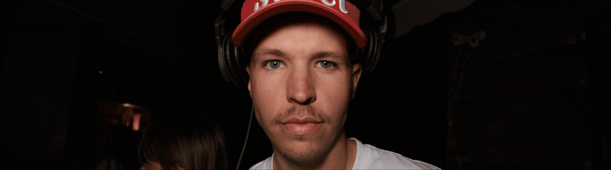 A young man wearing a red baseball cap and over-ear headphones, looking directly at the camera with a serious expression, in a dark environment.