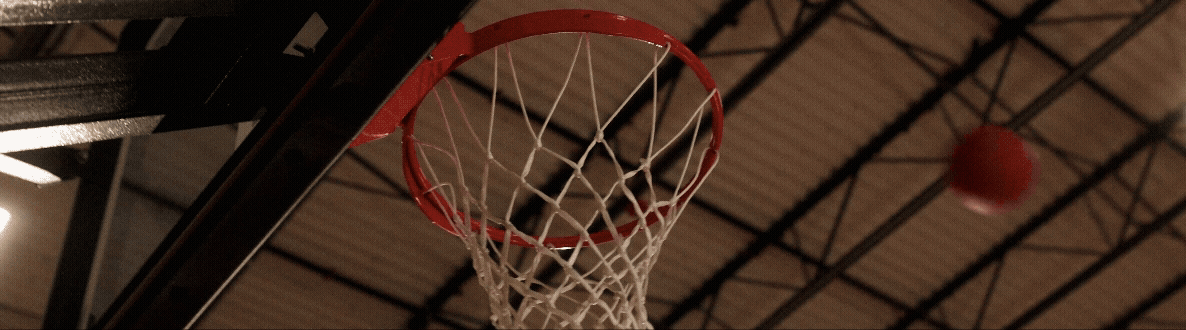 A basketball hoop with a net hanging from a metal rim, and a basketball approaching the hoop in an indoor gymnasium.