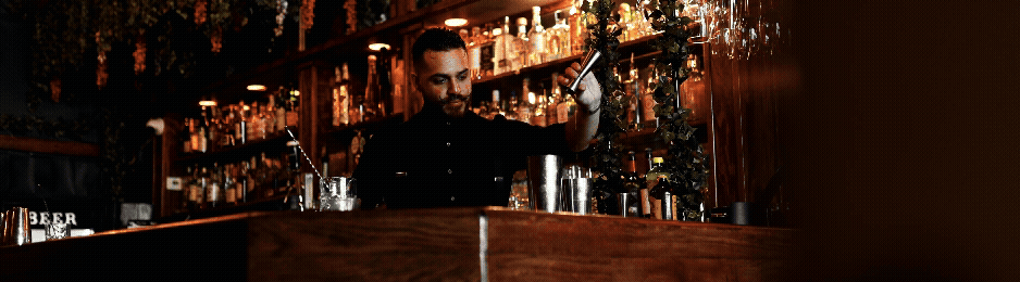 A bartender preparing a drink behind a dark wooden bar in a dimly lit bar or pub, with shelves of bottles in the background.