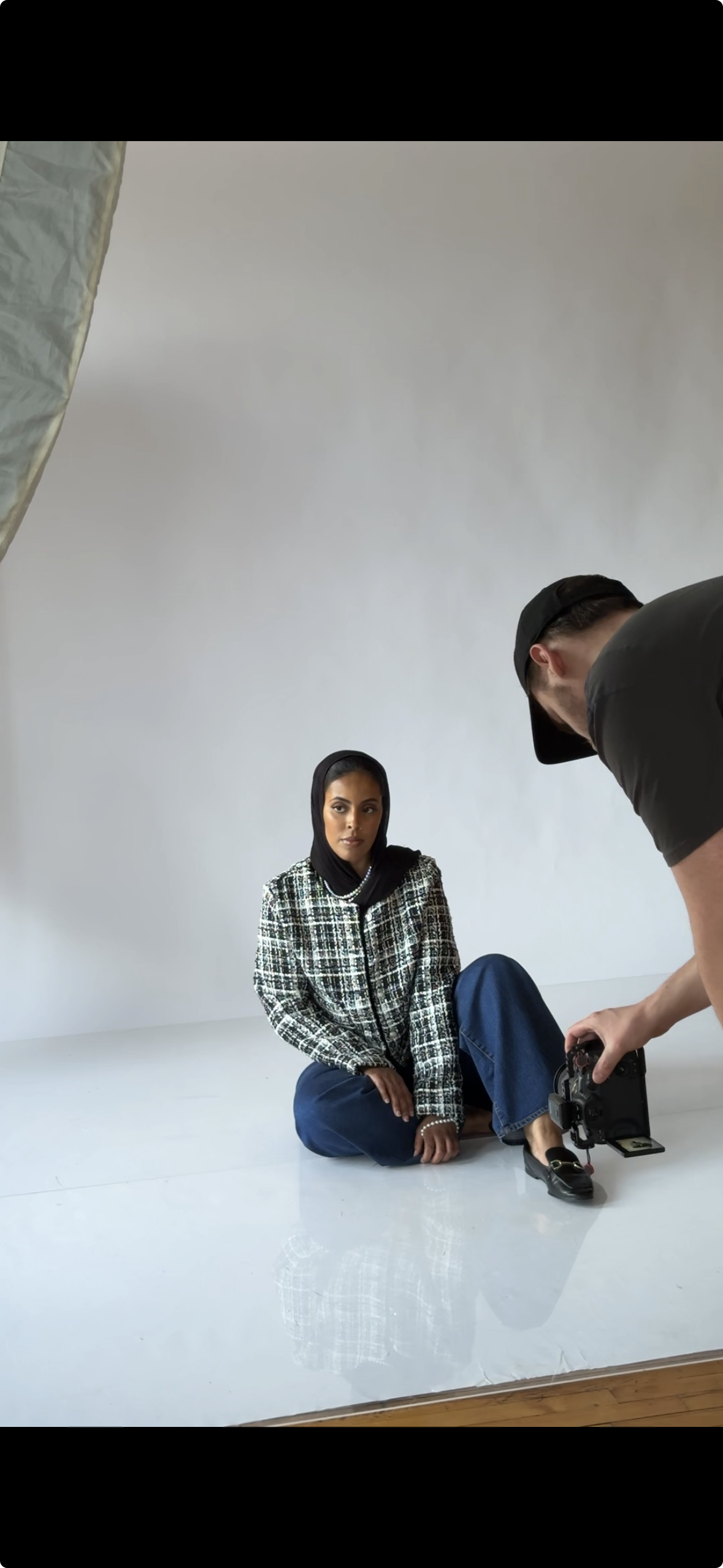 A woman with a black headscarf sitting on the floor for a photo shoot, with a photographer adjusting a camera nearby in a studio with a white backdrop.