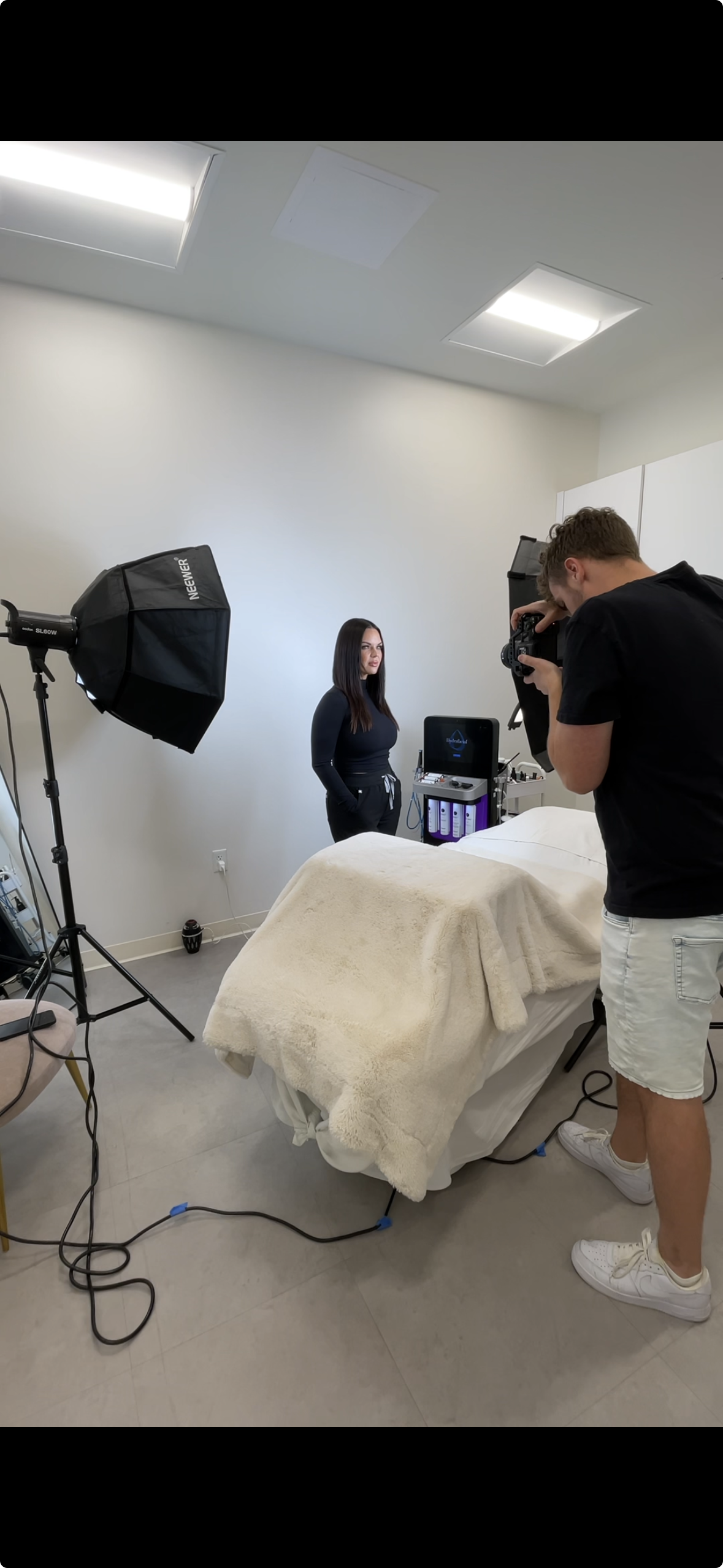 A woman standing in front of a white wall during a photo shoot or video recording, with a professional photographer taking pictures. The scene includes studio lighting, a bed with a beige blanket, and some medical or skincare equipment in the backgro