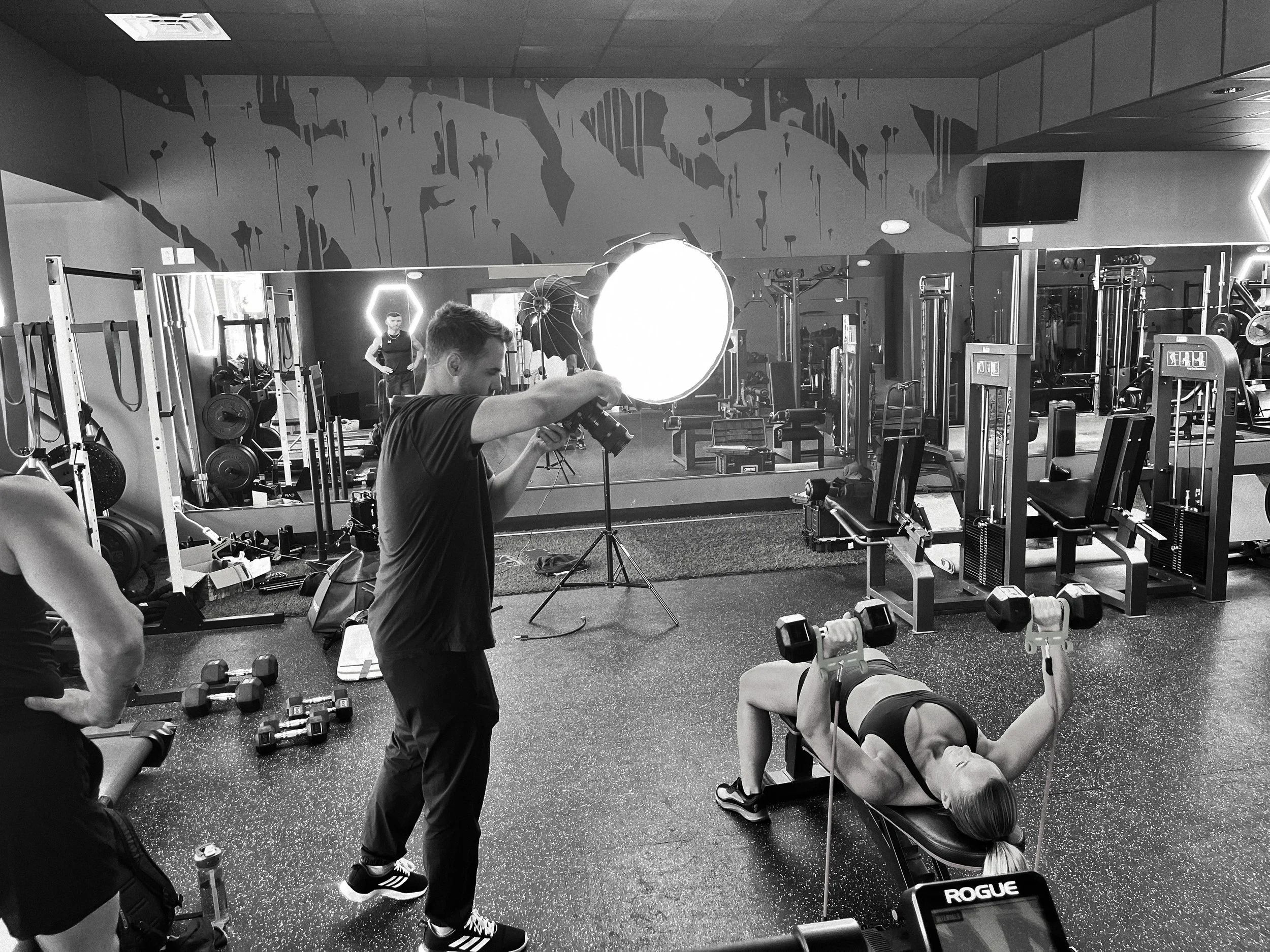 Photograph of a gym workout scene in black and white. A woman is lying on a workout bench lifting dumbbells, with a camera crew filming her using a large light. Other gym equipment, weights, and a person adjusting their hair can be seen in the backgr