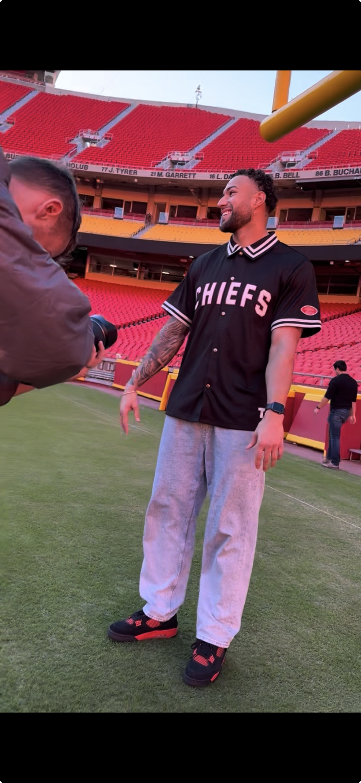 A man in a black Chiefs baseball jersey and gray sweatpants standing on a football field, being photographed by a person holding a camera. The stadium seats are visible in the background.