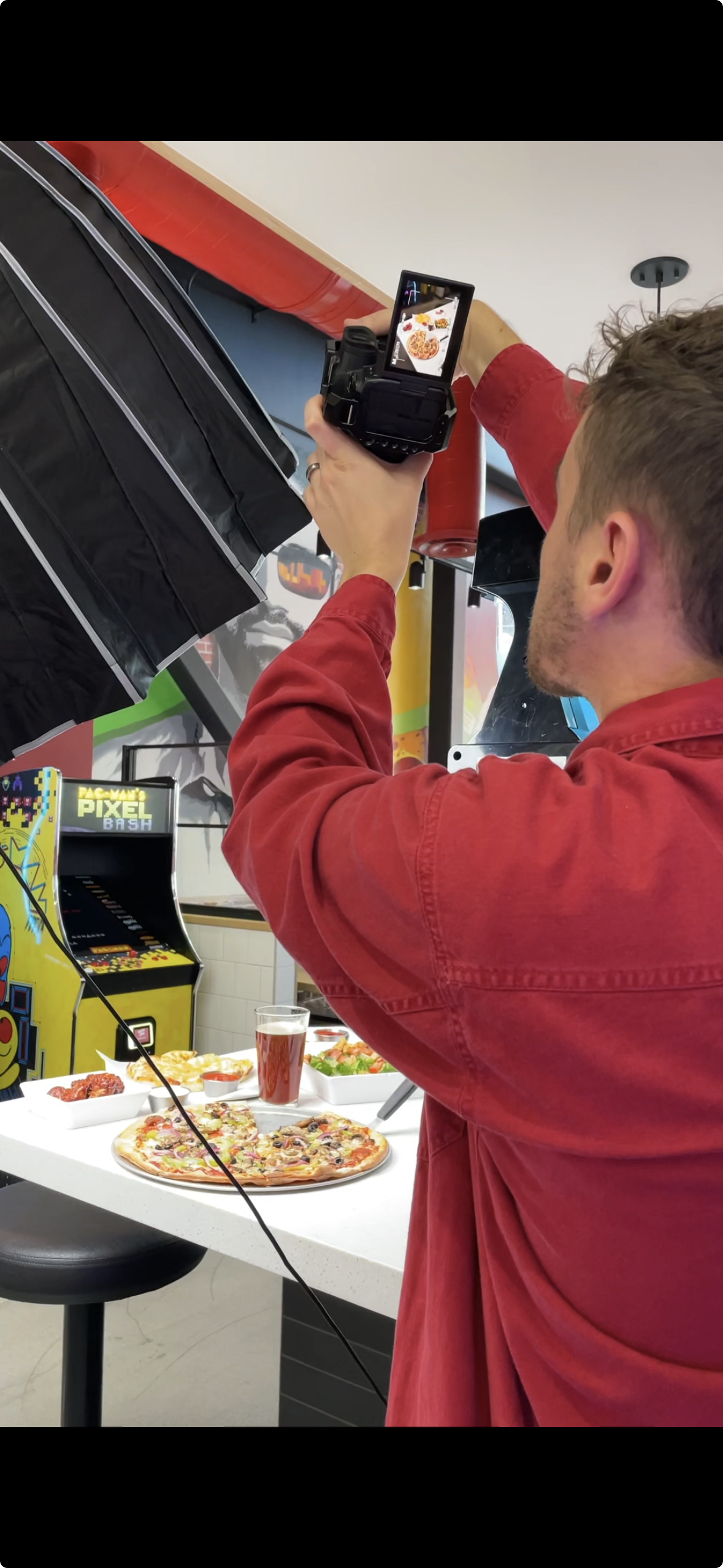 A man in a red shirt taking a photo of pizza and snacks on a counter with a camera in a room with arcade games.