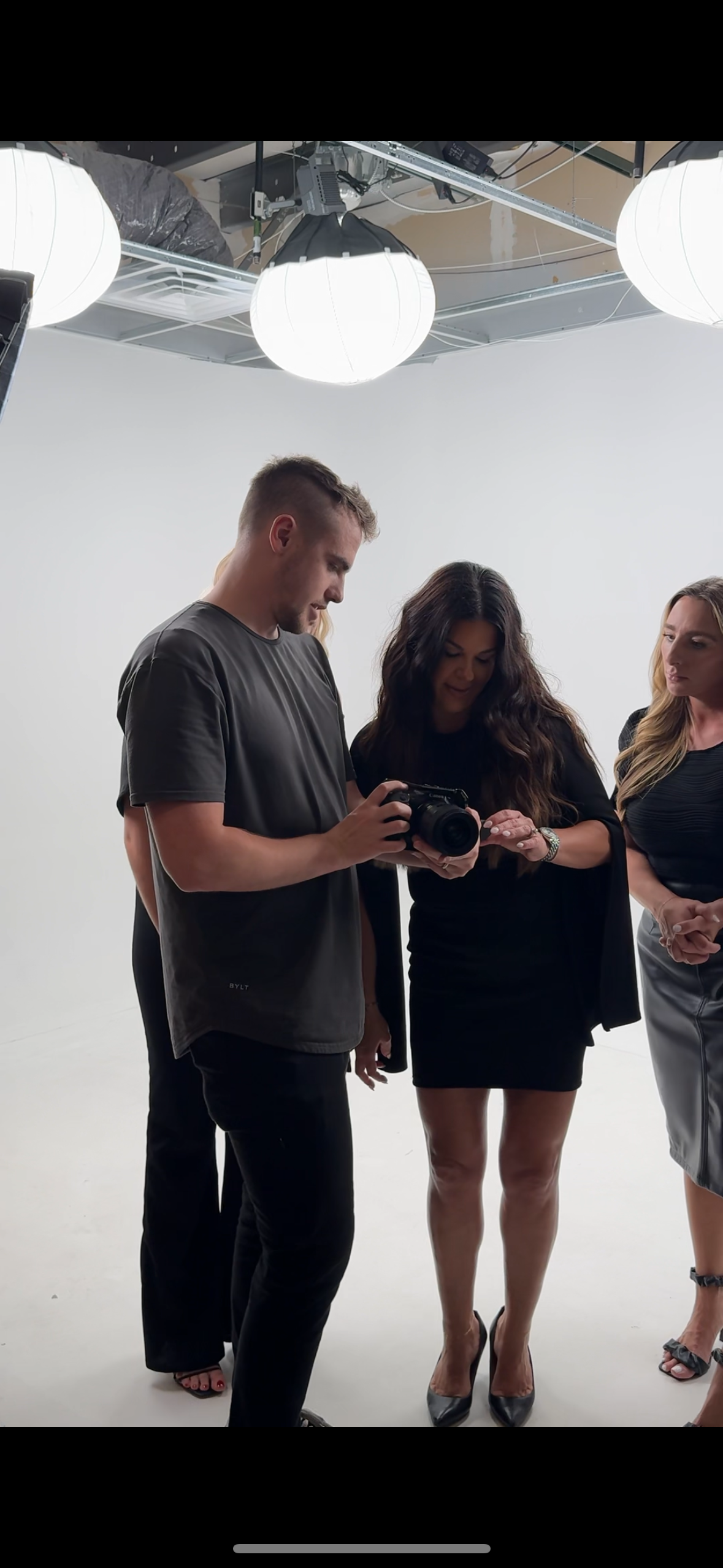 Three people standing together in a studio with a plain white background, two women and one man, looking at a camera that the man is holding, under bright studio lights.
