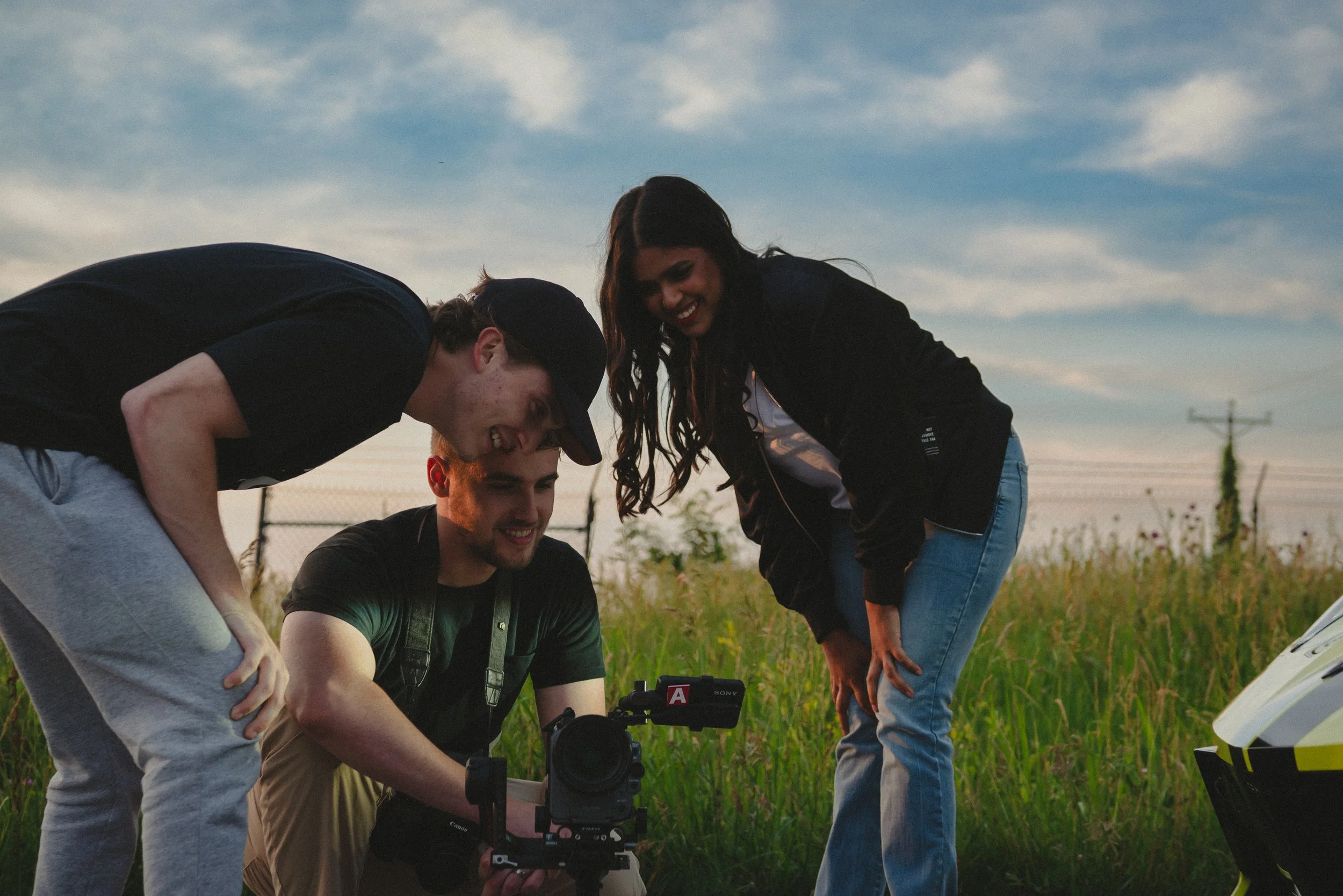 Three young adults gathered around a camera on a grassy field at sunset, smiling and looking at the camera's viewfinder. Two men and one woman appear to be filming or taking photos, with a motorcycle helmet visible on the right.