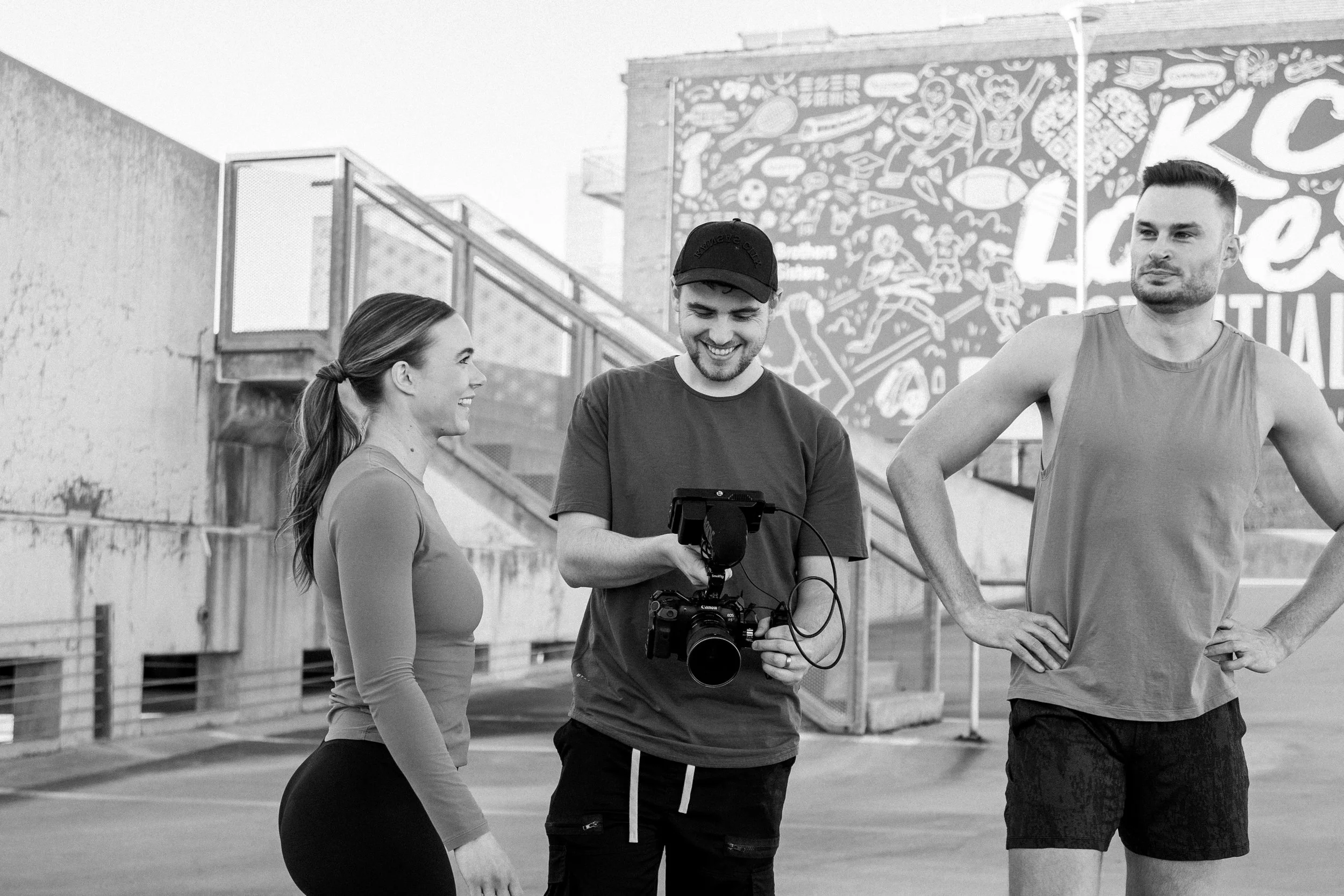 A woman, a man with a camera, and a man in a sleeveless shirt standing outdoors, smiling and talking.