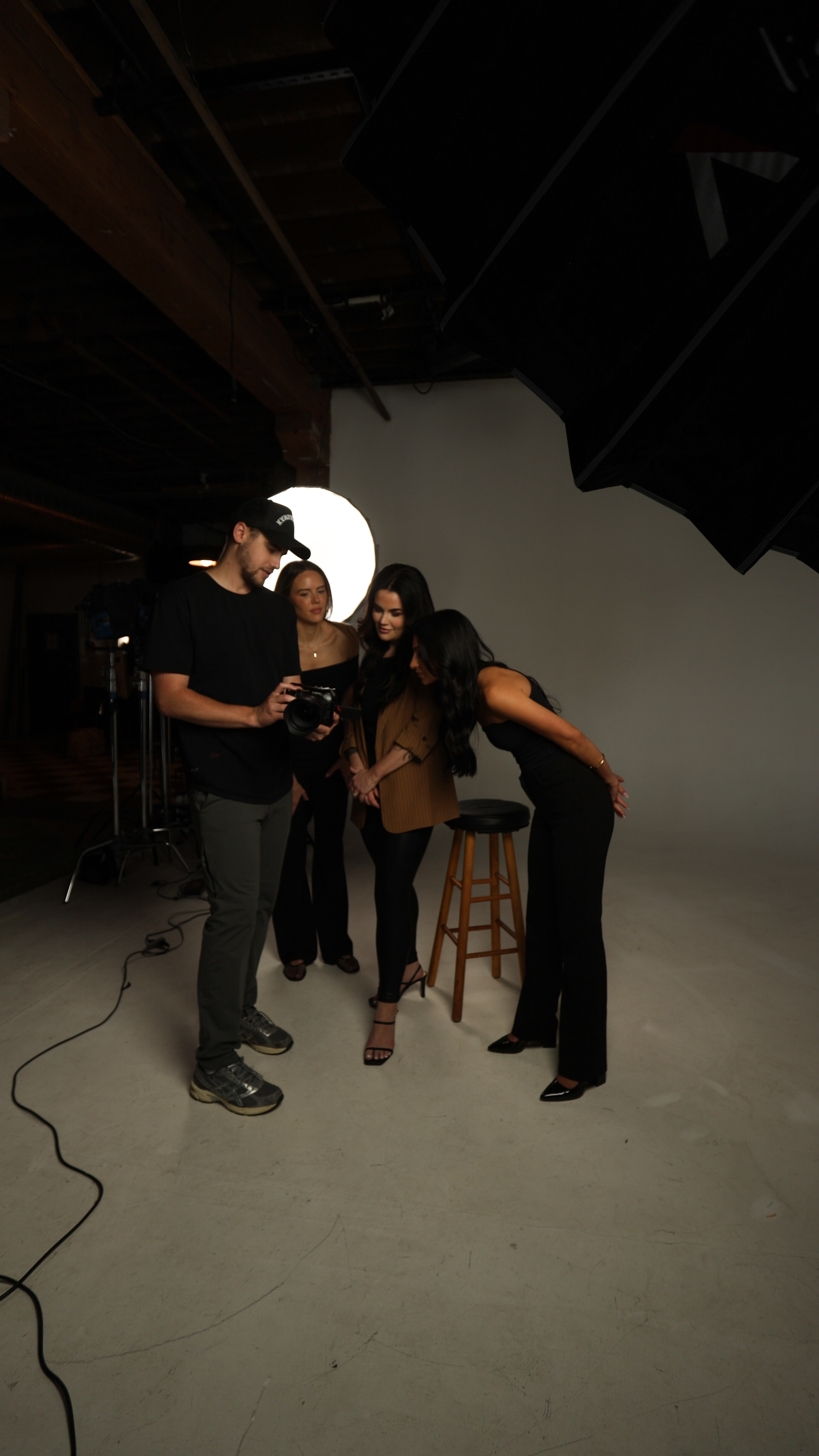 Four people in a photography studio looking at a camera, two women and one man mostly in black and one woman in a brown jacket. A photography lighting setup is visible in the background.