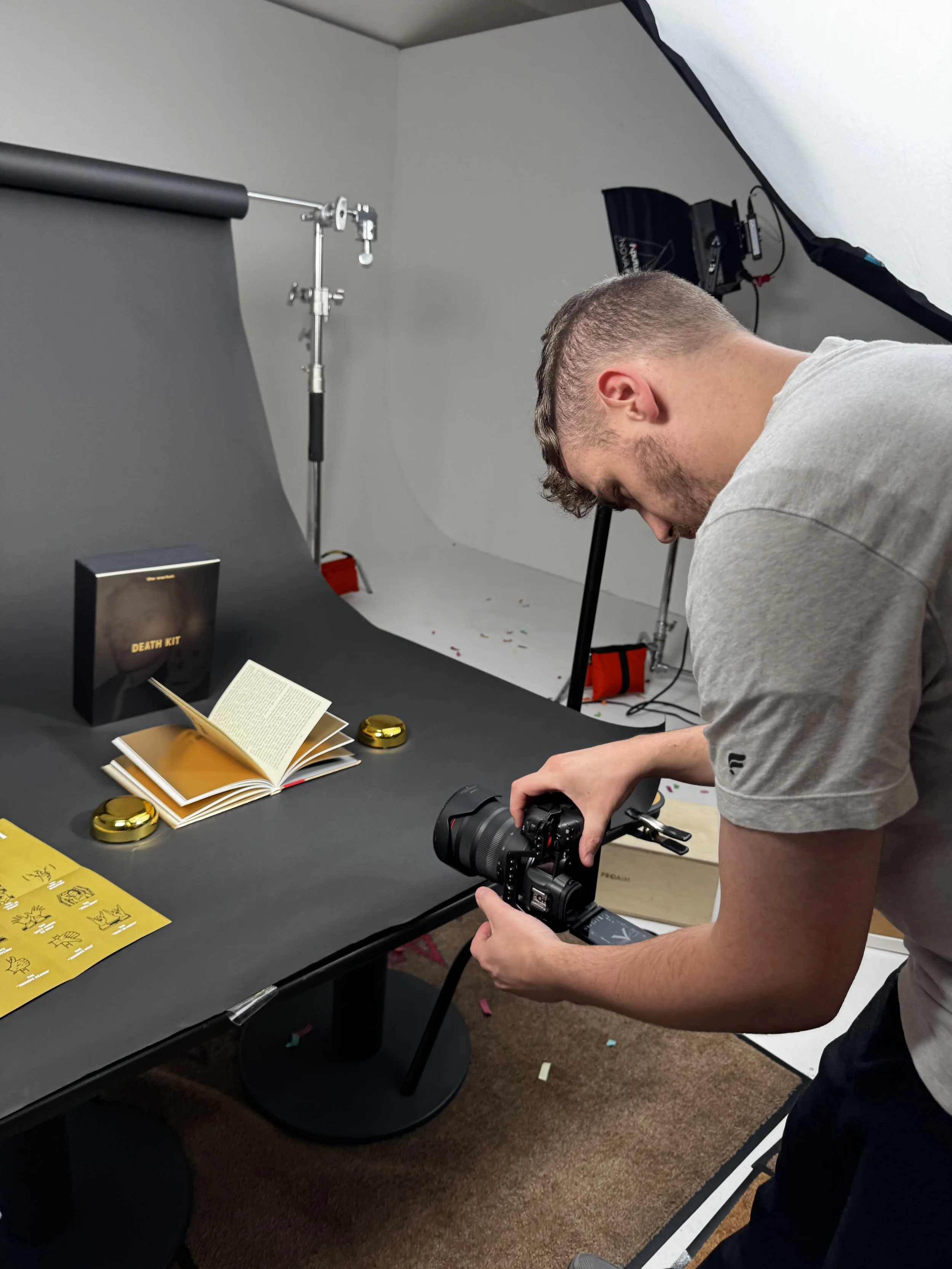 A man with a gray T-shirt is setting up a camera on a Dick Blick black table with a black background paper, writing and gold compact cases, in a photography studio with a white backdrop and professional lighting.