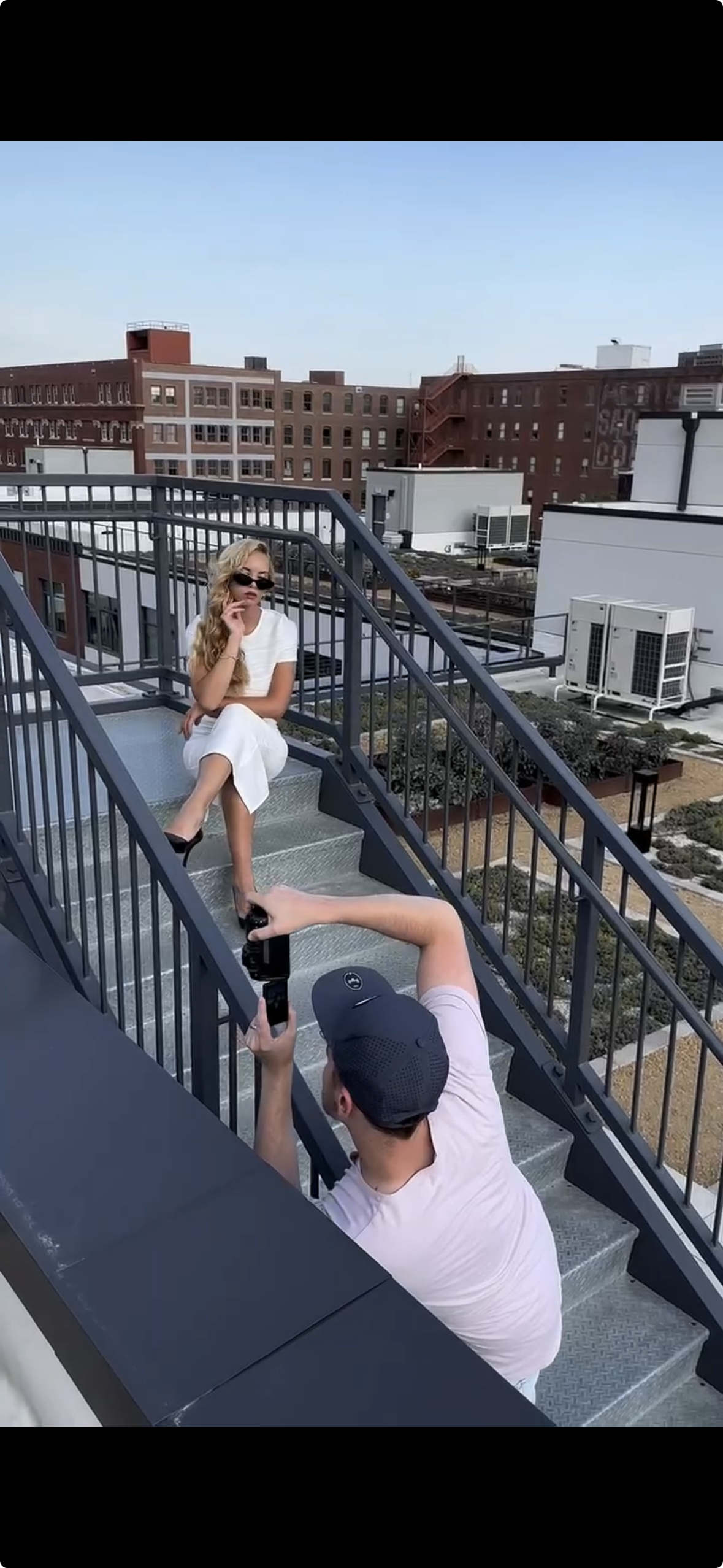 A woman in a white dress is sitting on a rooftop staircase, posing for a photo taken by a man in a light-colored shirt and baseball cap, with city buildings in the background.