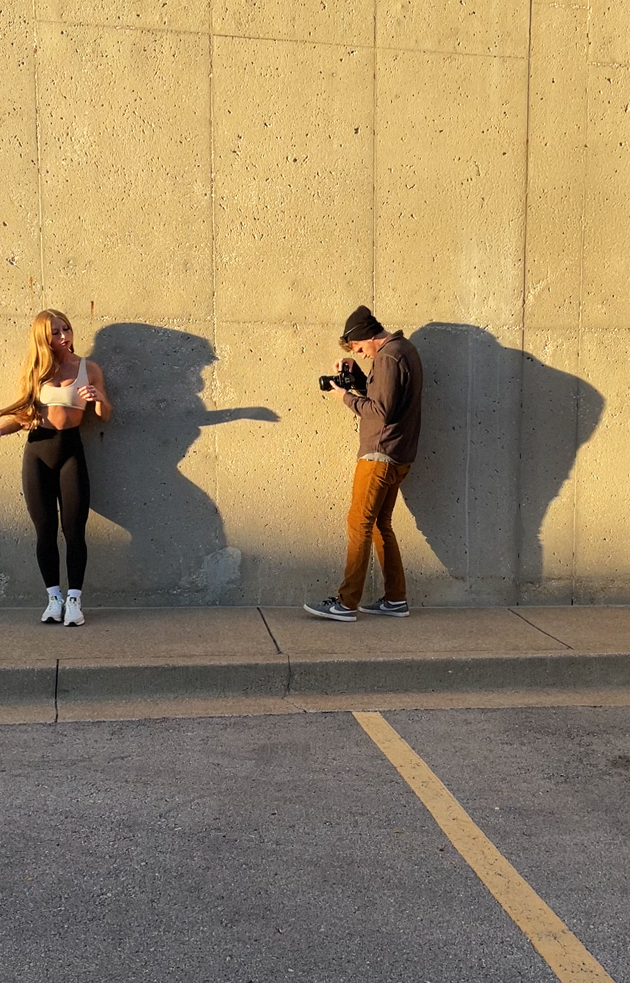 A woman in athletic clothing standing against a concrete wall as a man with a camera takes her picture during sunset, casting shadows on the wall.