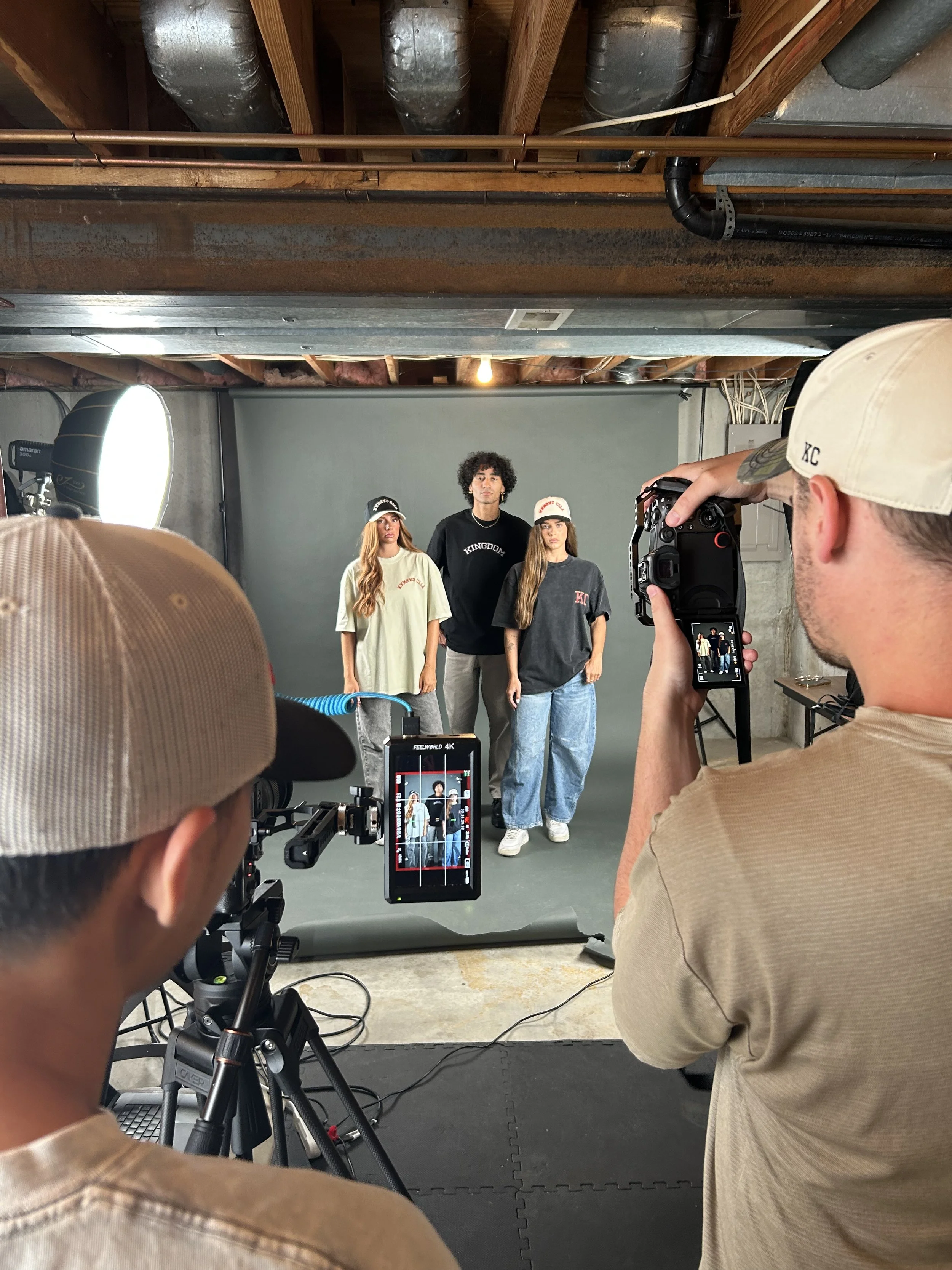 People behind the camera taking a picture of three models standing in front of a gray backdrop in a photography studio.