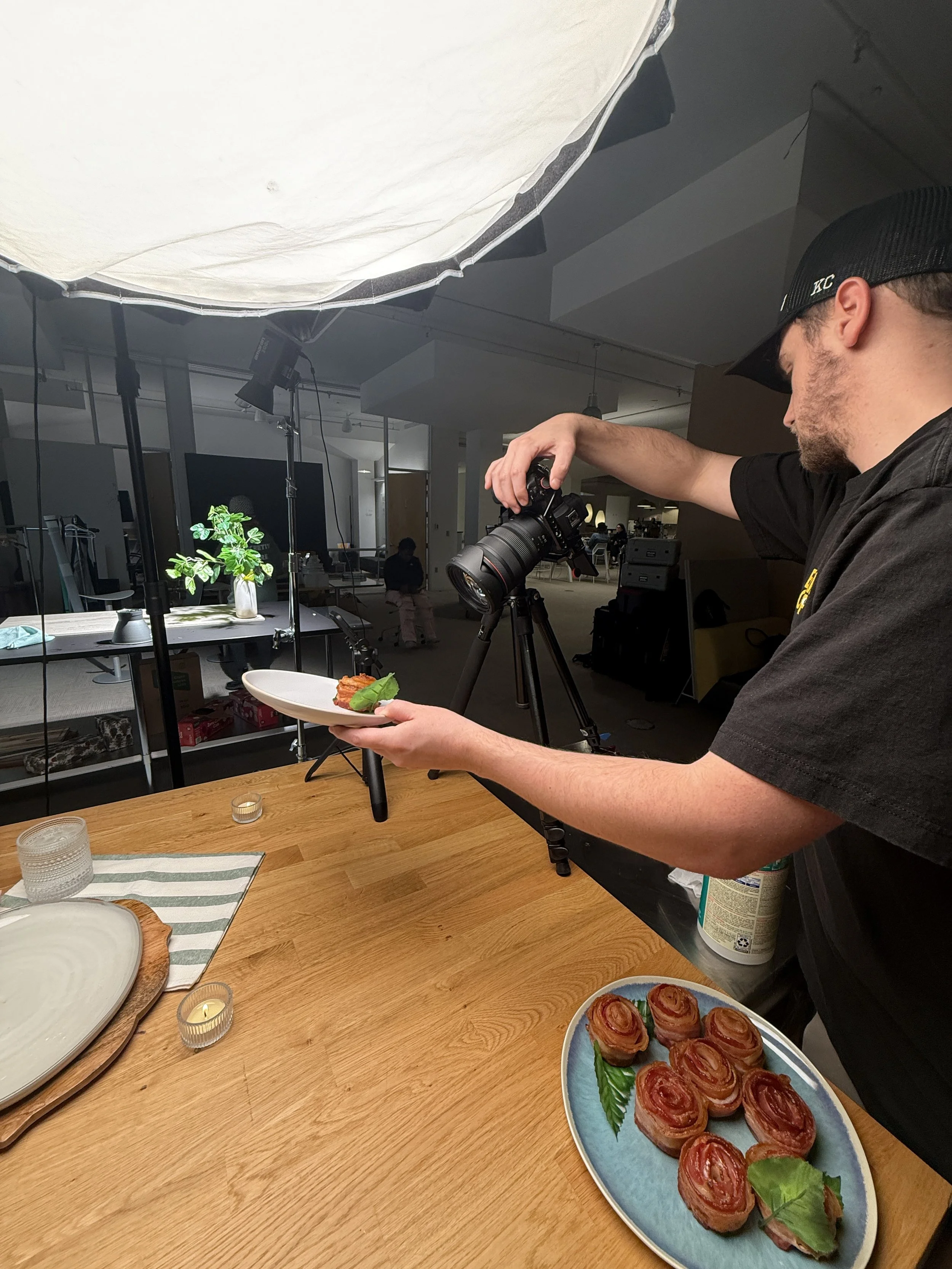 A man wearing a black cap and shirt is setting up a food photograph with a camera on a tripod. The man holds a small plate with food, while a plate of rolled bacon-wrapped appetizers with basil leaves is placed on the wooden table. A large softbox li