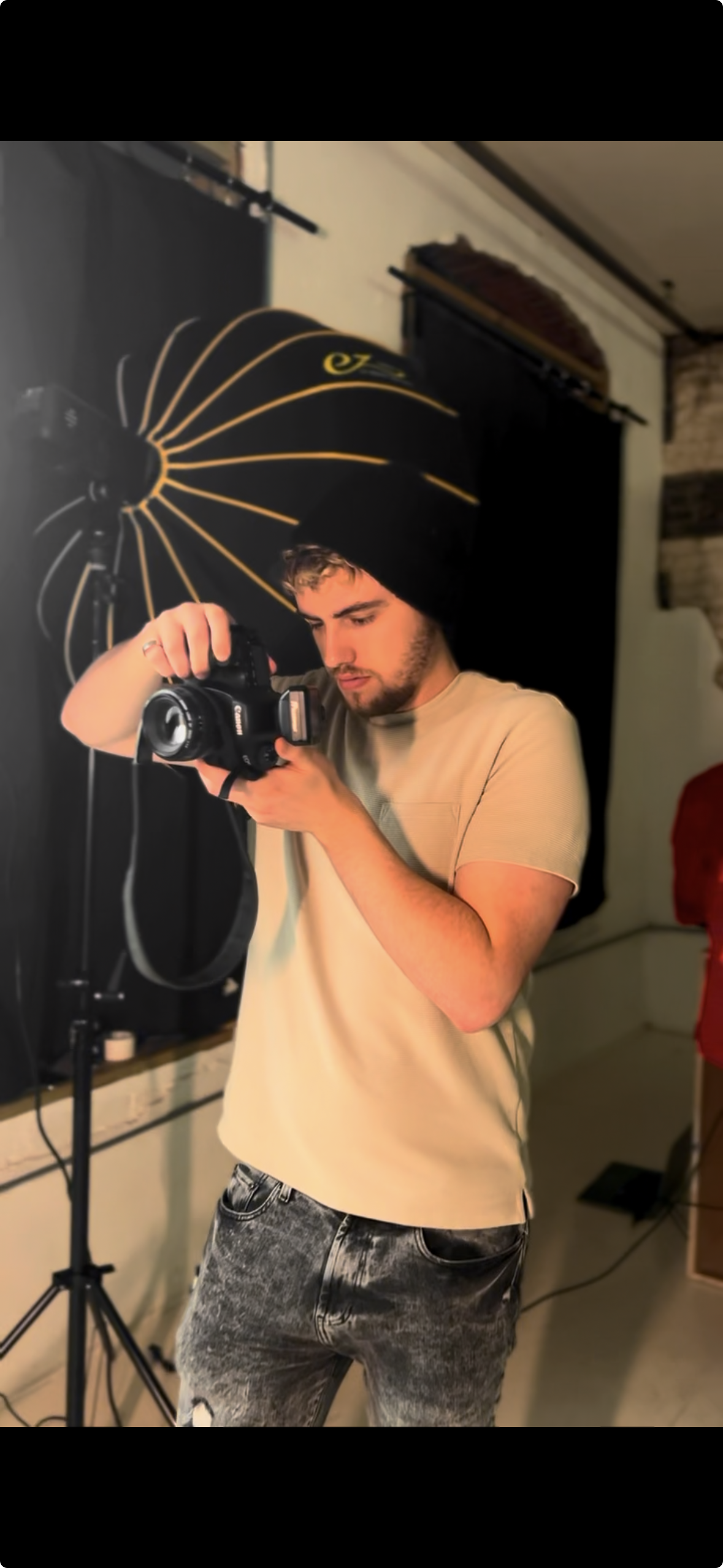 Young man in beige T-shirt and black jeans taking a photograph with a DSLR camera in a photography studio with lighting equipment and backdrop.