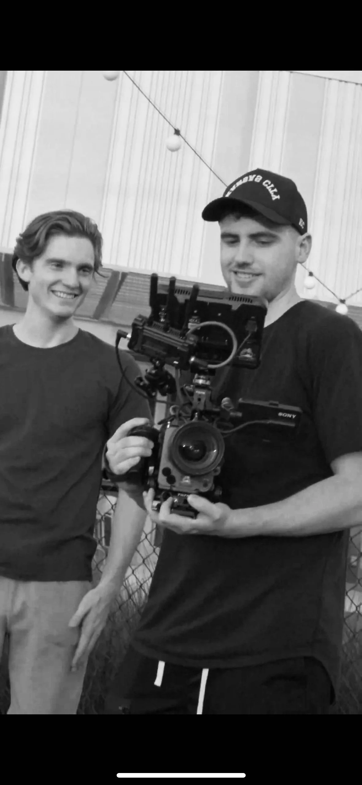 Two young men, one smiling, are having a photo taken together. One is holding a professional camera with a stabilizer, and the other is standing nearby.