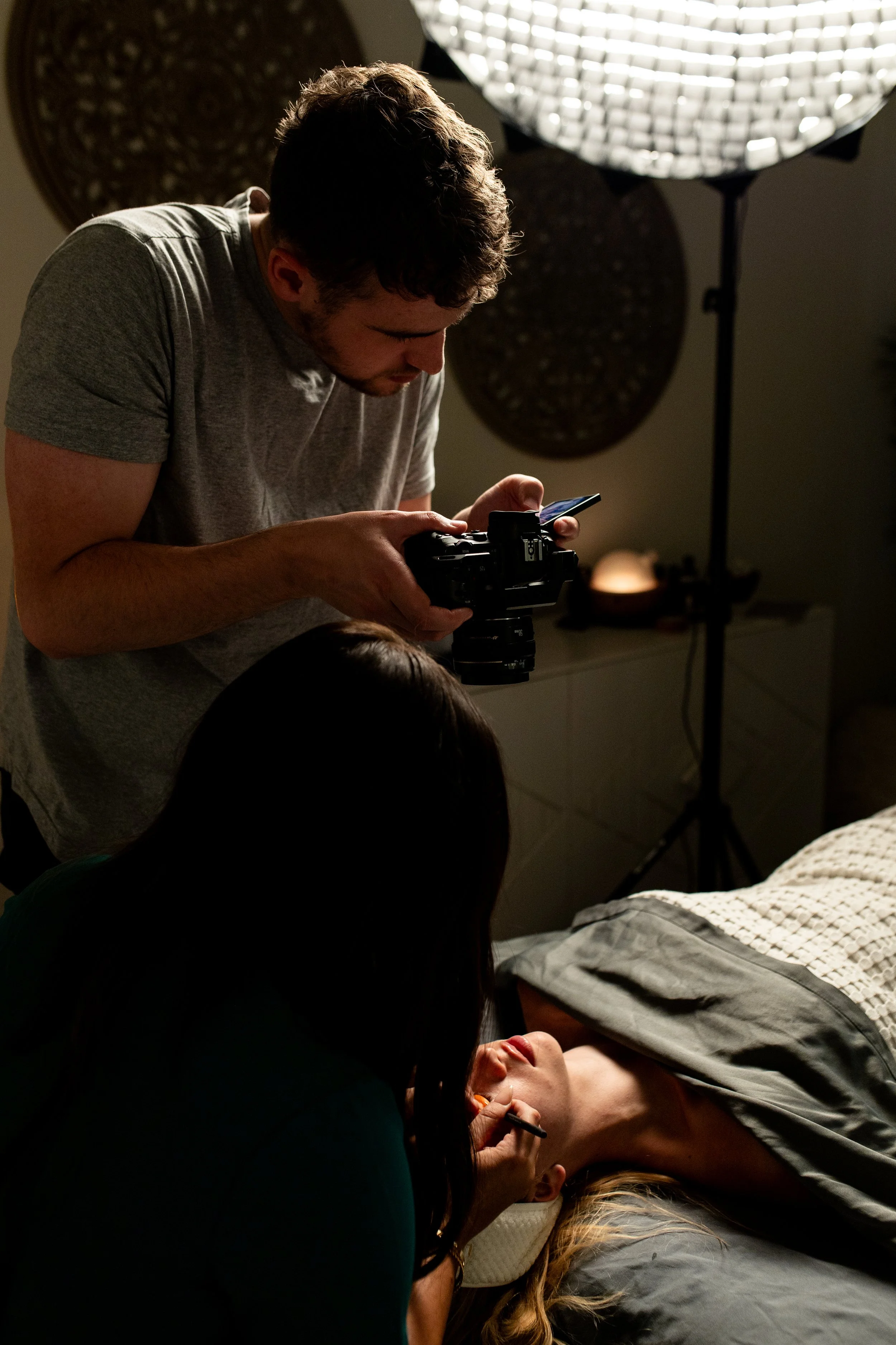 A photographer reviews a shot on a camera as a makeup artist applies makeup to a woman lying on a bed in a dimly lit room with professional lighting equipment.