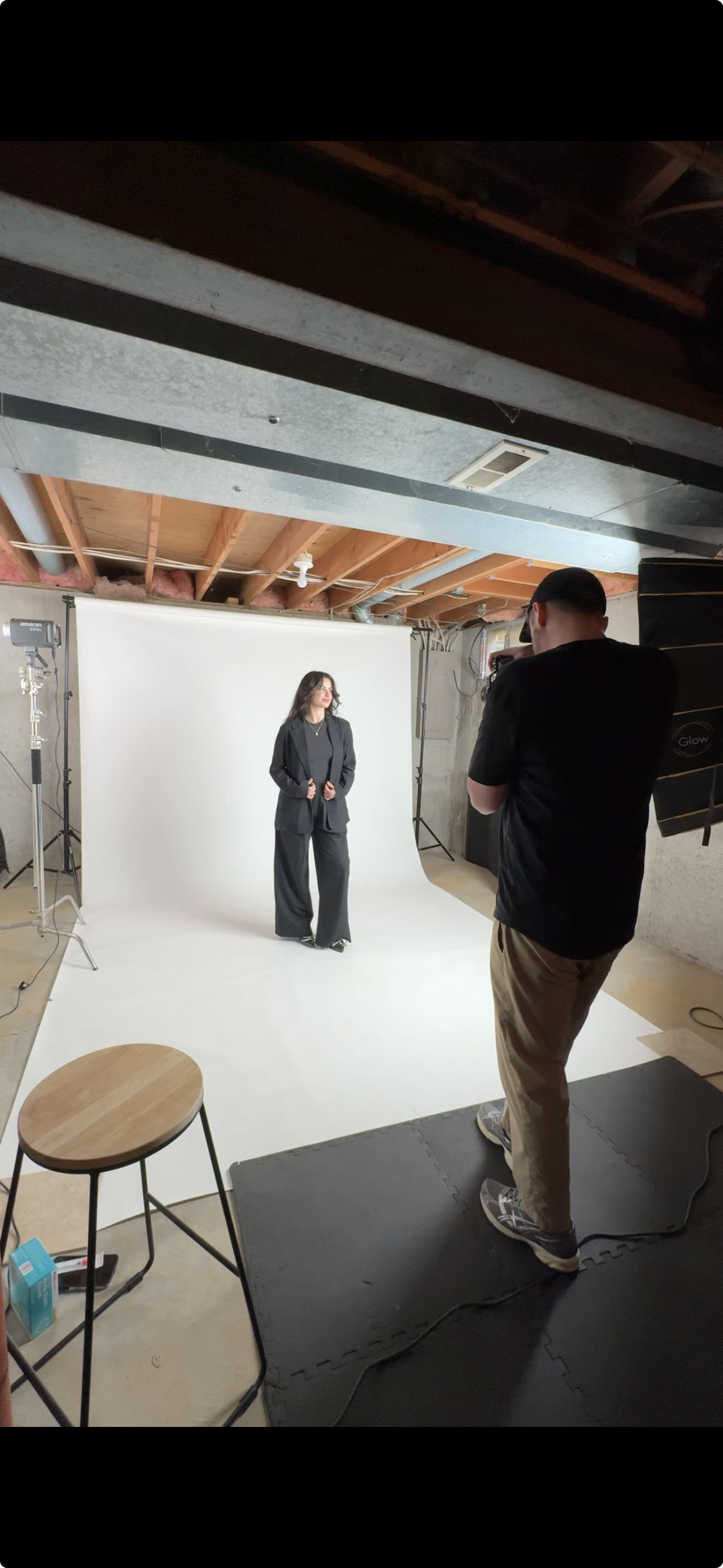 A woman in a black suit stands on a white backdrop in a photography studio, while a male photographer captures her photo.