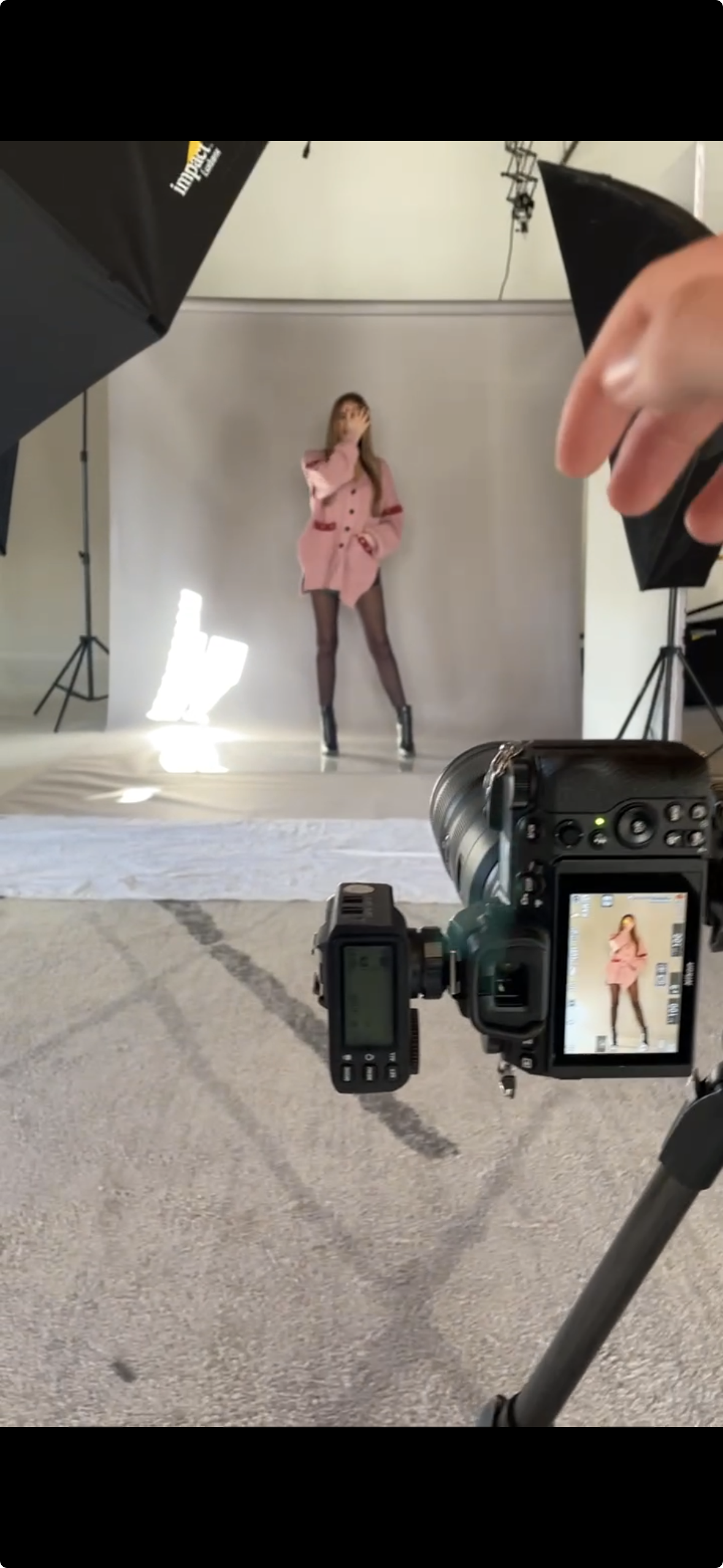 A woman in a pink coat and black tights posing in front of a grey backdrop during a photo shoot, with photography equipment including lights and a camera capturing her image.