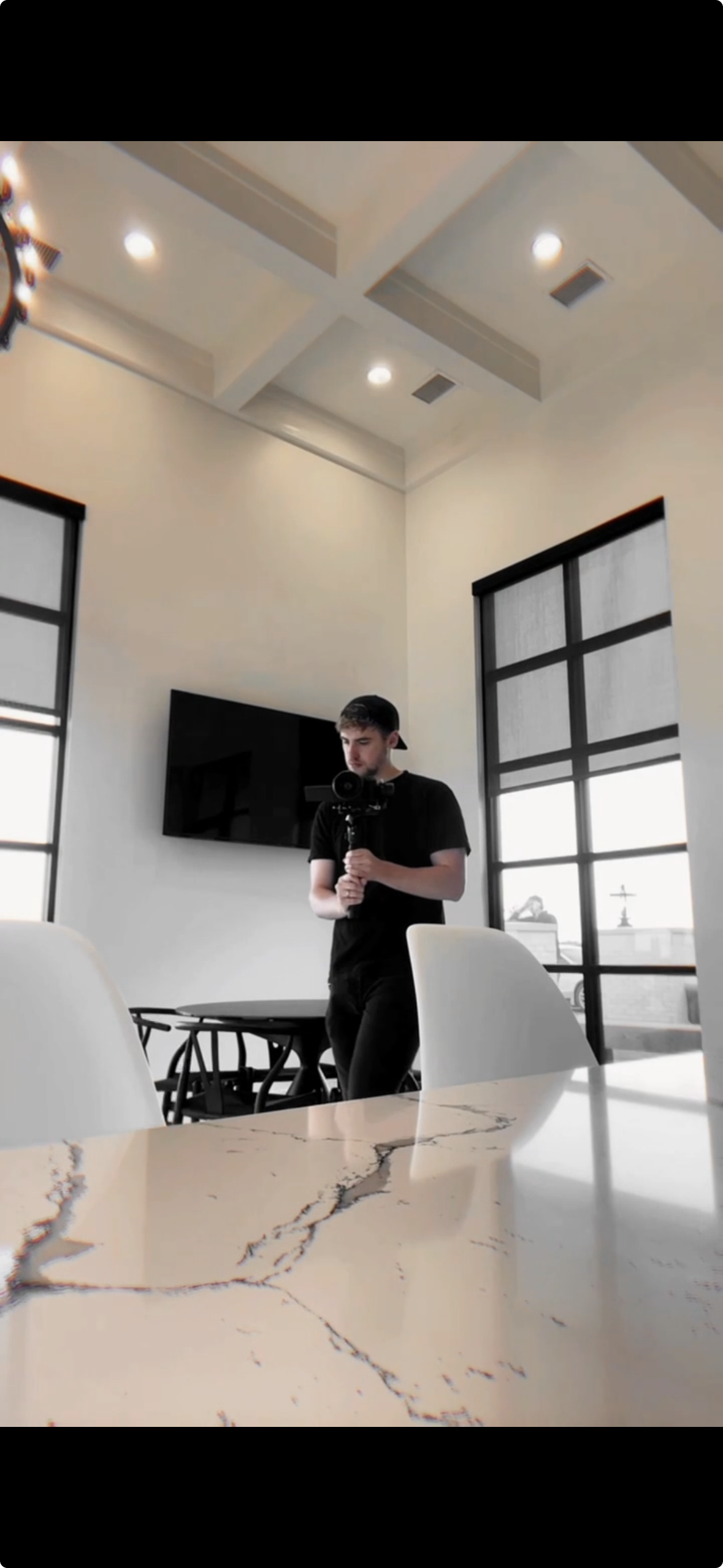 Young man in black shirt and cap taking a selfie with a camera in a modern, bright room with large windows, a marble table, and a wall-mounted TV.