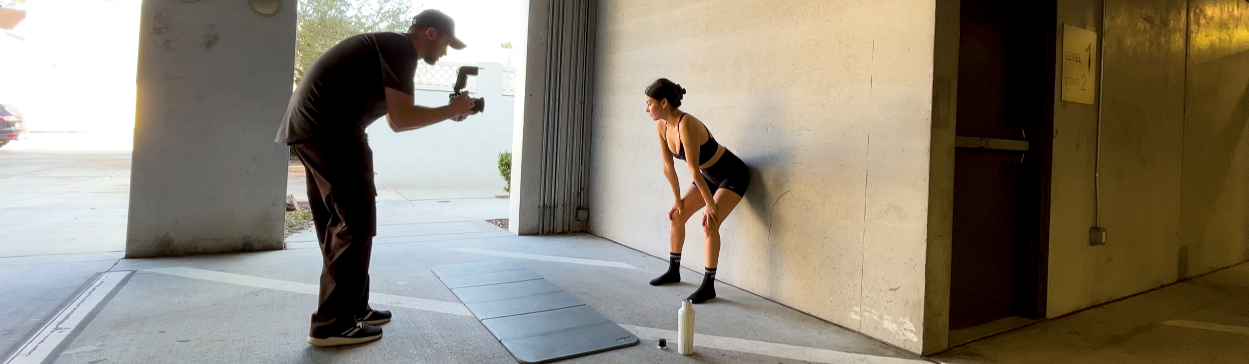 A woman in sporty attire stands with her hands on her knees, leaning against a wall, as a man records her with a camera inside a parking garage or similar structure.