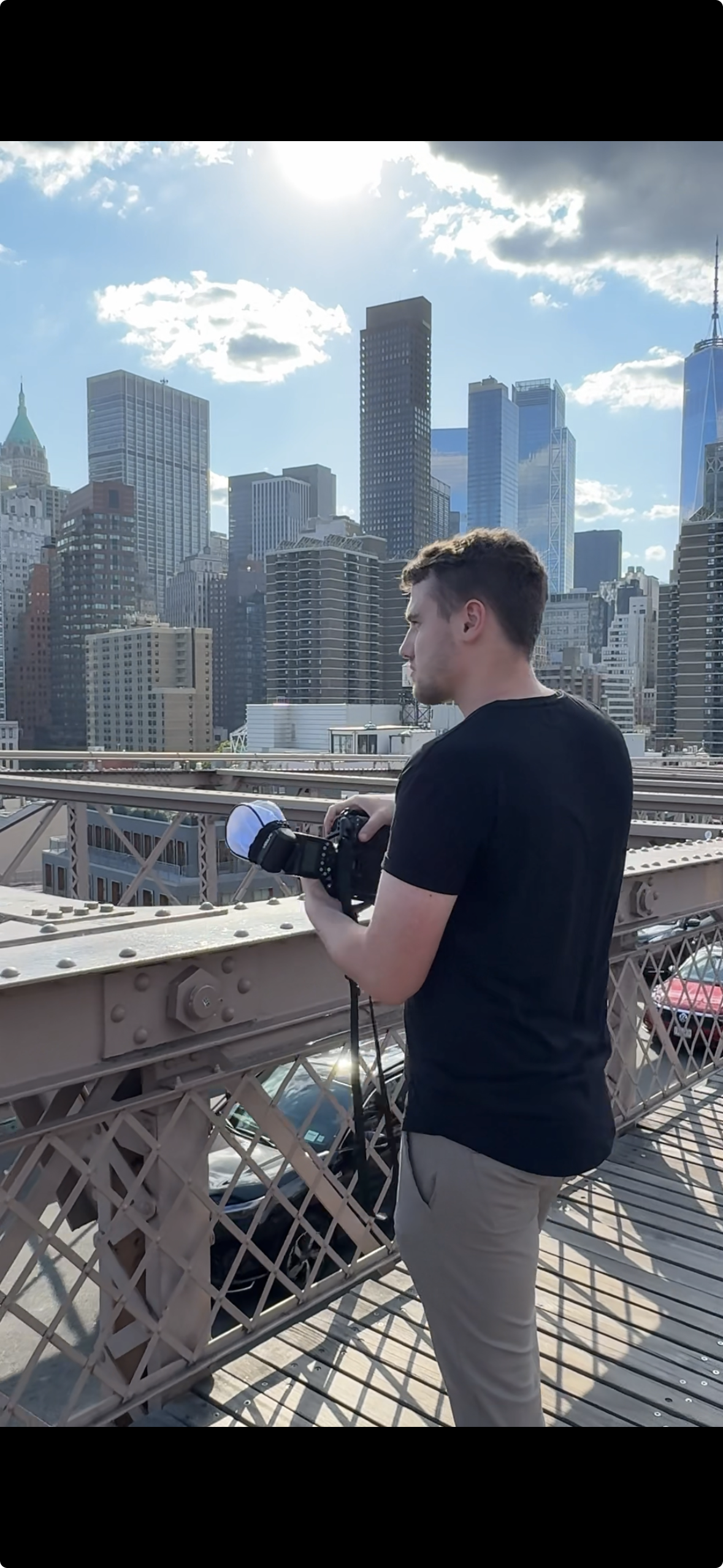 Young man standing on the Brooklyn Bridge with a camera, overlooking the New York City skyline with tall buildings under a partly cloudy sky.