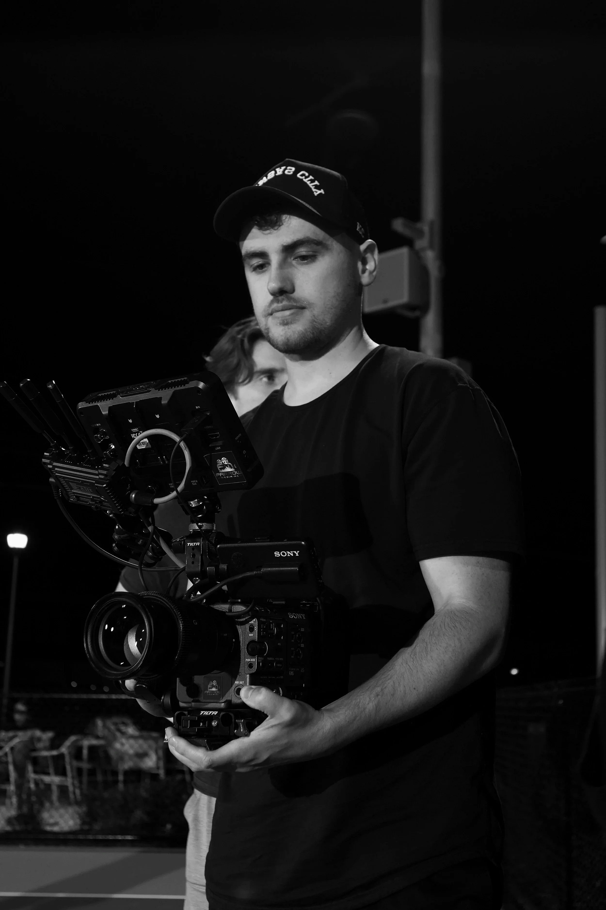 Young man holding a professional camera equipment setup during nighttime, with another person partially visible behind him.