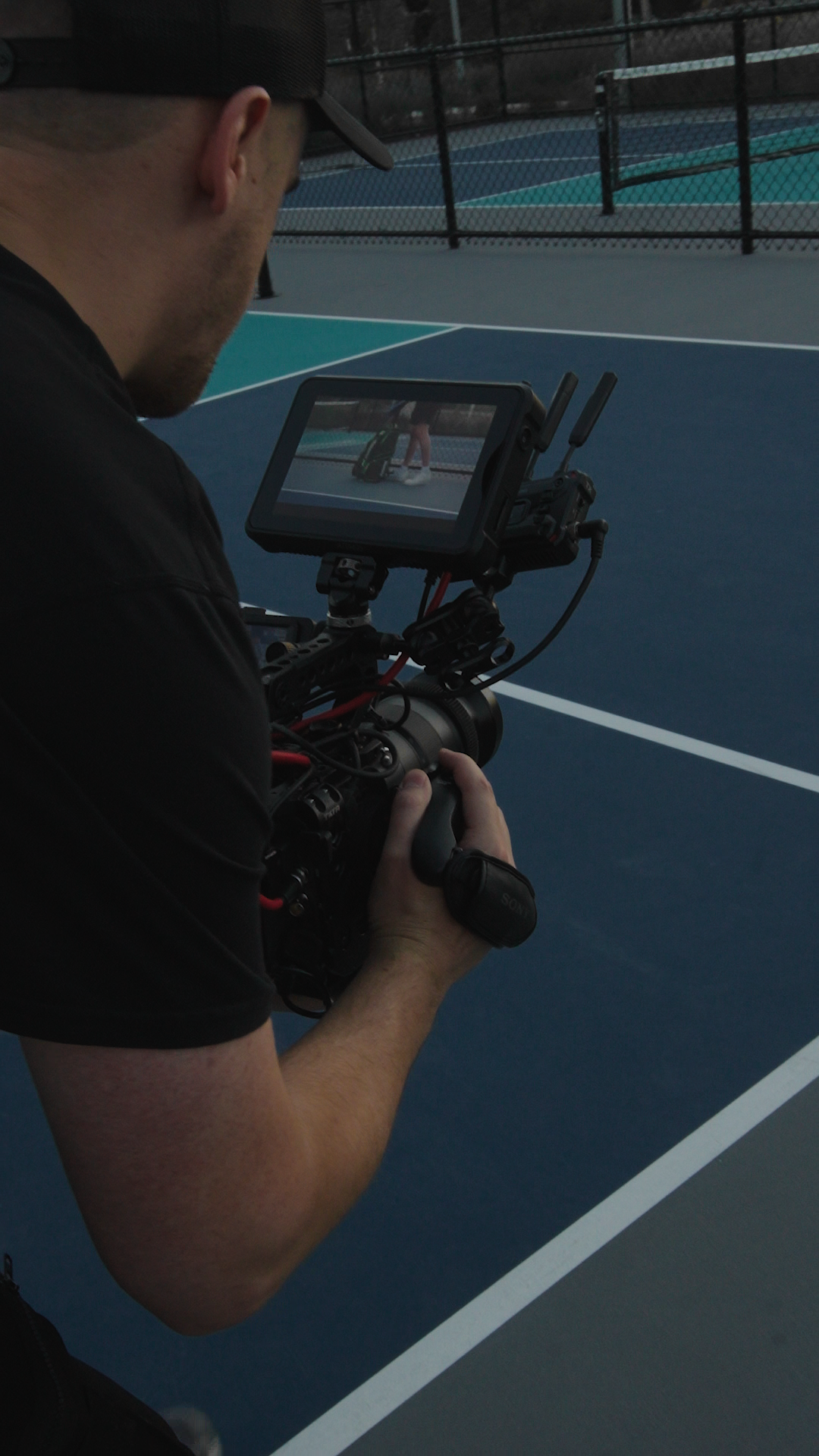 A person operating a professional video camera on a tennis court, filming a person preparing to serve.
