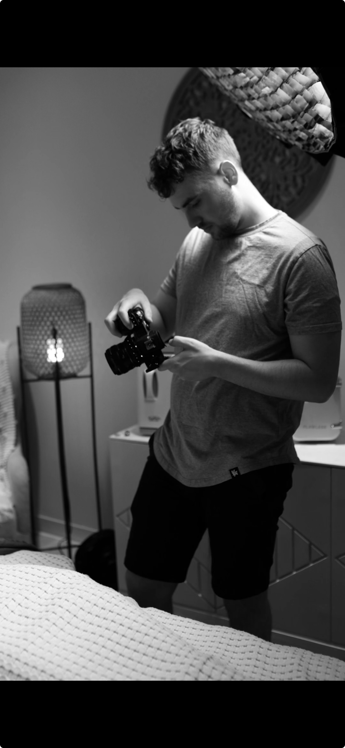 A young man with curly hair wearing a t-shirt and shorts is holding a camera, looking down at it, in a room with a bed, lamp, and dresser.
