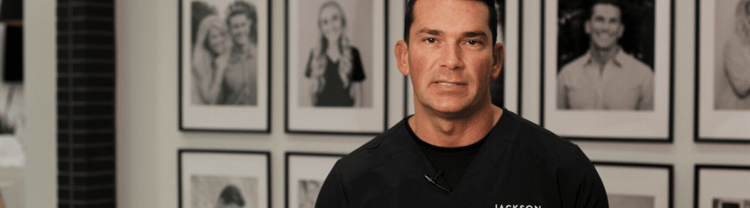 A man in a black shirt stands in front of a wall with framed black-and-white photographs of various people.