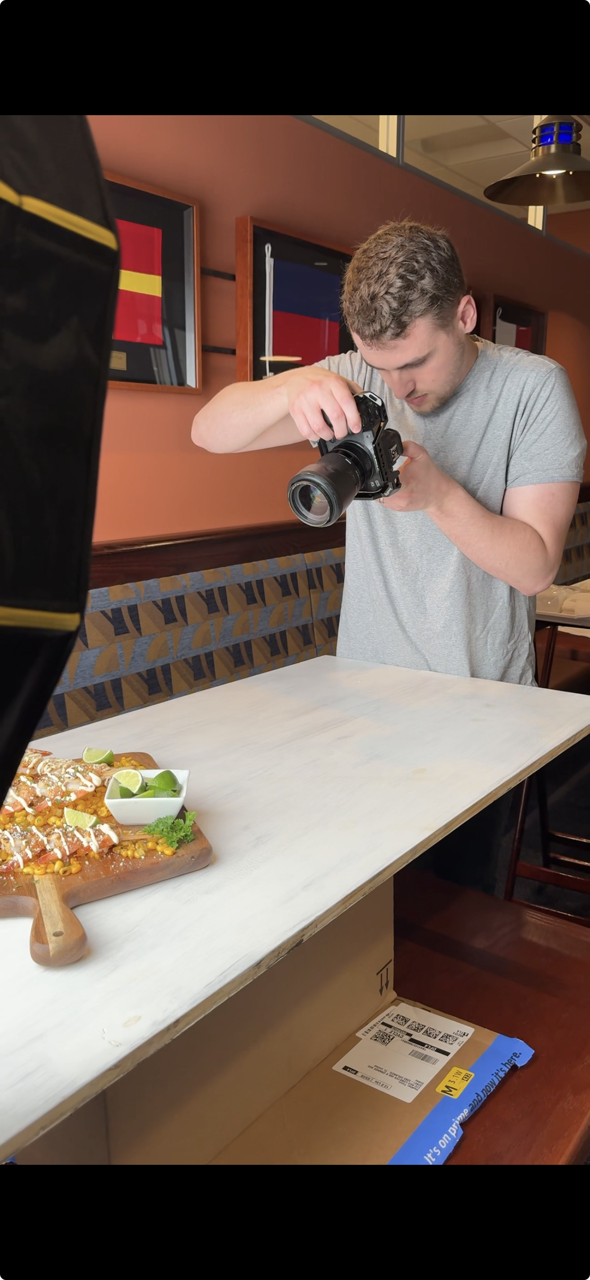 A young man with curly hair wearing a gray t-shirt is taking a photo of a food dish with a professional camera inside a restaurant. The food on the dark wooden serving board includes sliced fish, corn, lime wedges, and garnishes. The background featu