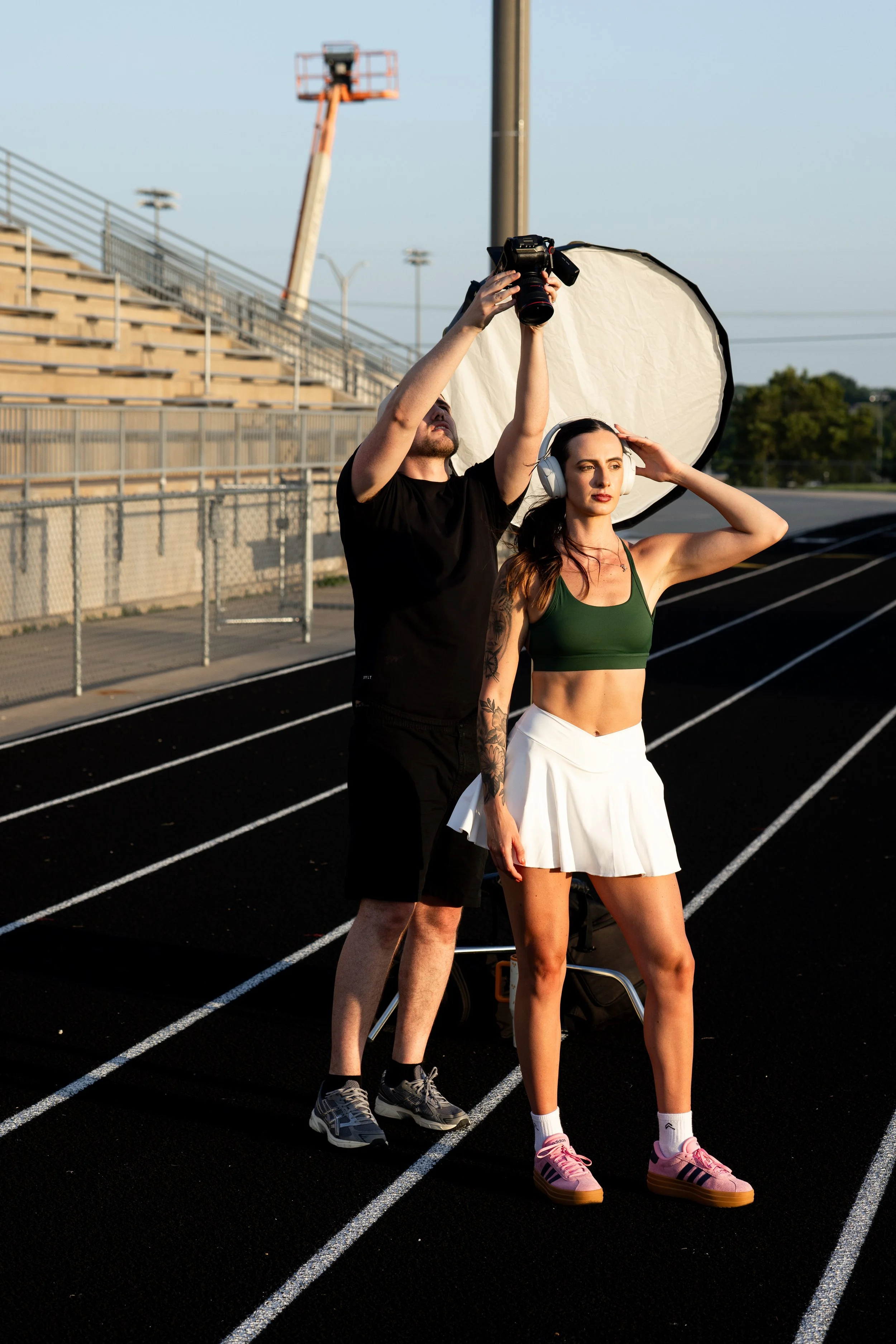 A woman in athletic clothing, headphones, and pink sneakers poses on a track while a photographer adjusts a lighting reflector and camera during a photoshoot.