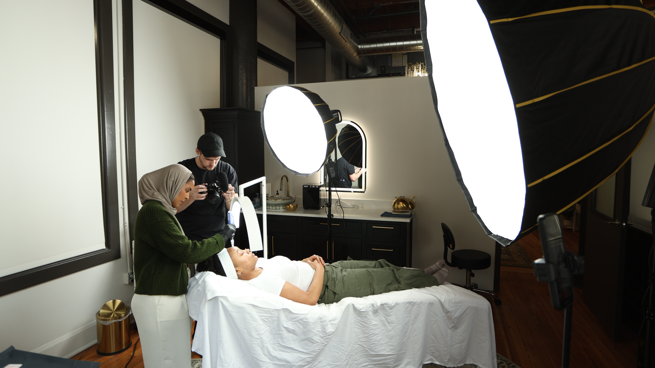 Photo of a woman lying on a bed with her eyes closed, surrounded by two people using professional studio lighting and cameras, in a room set up for a photography session.