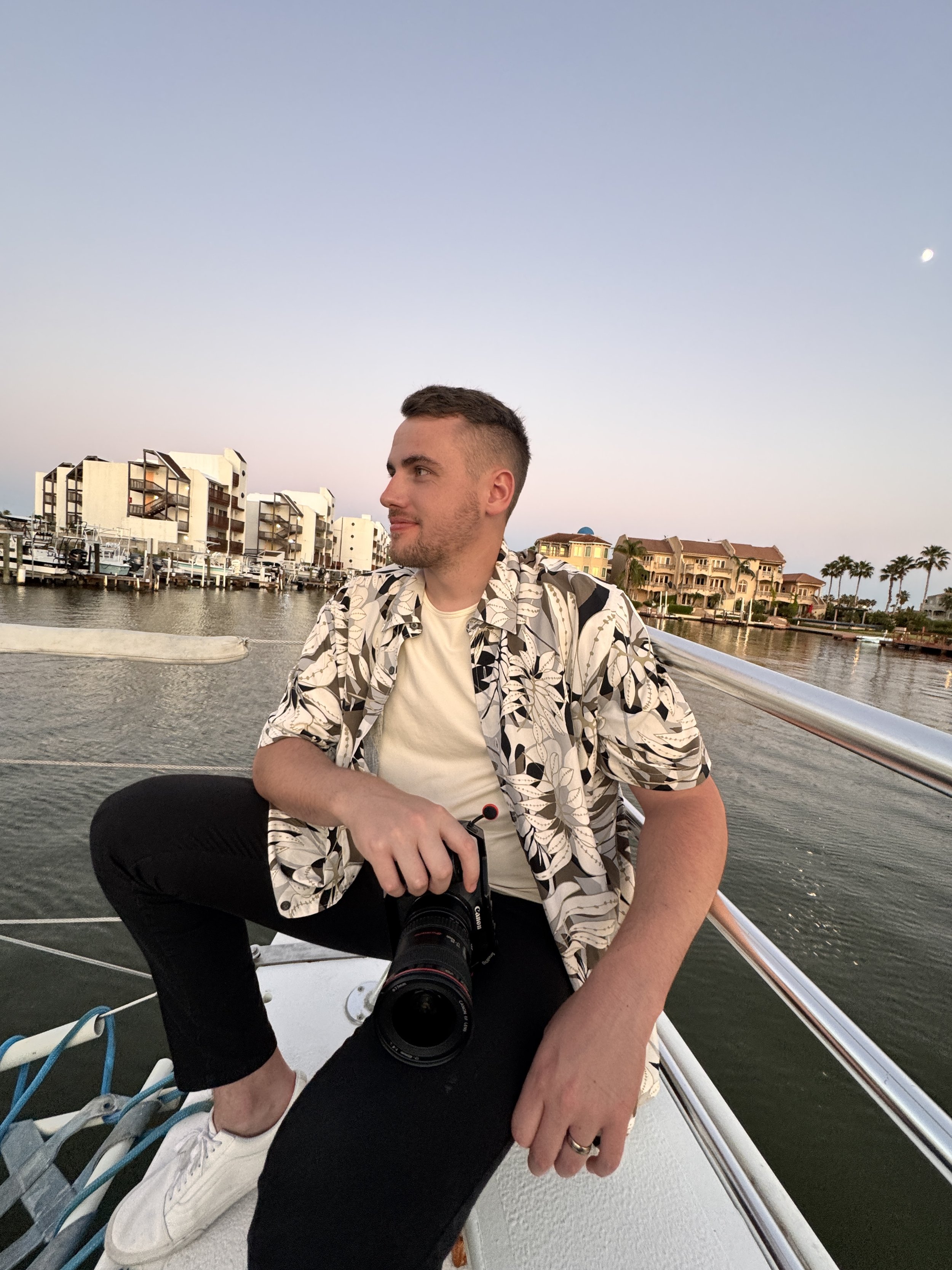 A man sitting on the edge of a boat with a camera in hand, overlooking a marina with houses and palm trees at sunset.